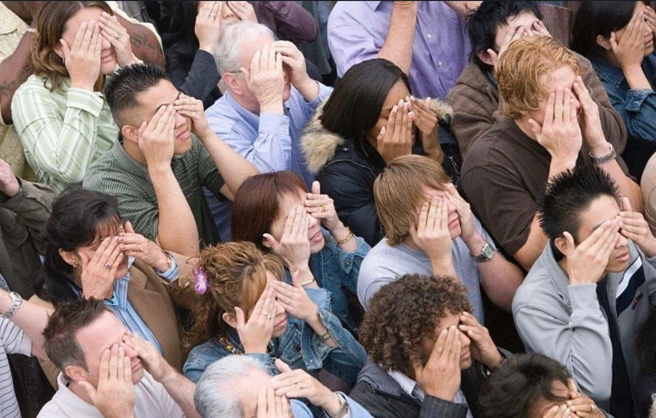 A group of people are sitting together with their eyes closed and hands covering their faces, participating in a shared moment of reflection or meditation.