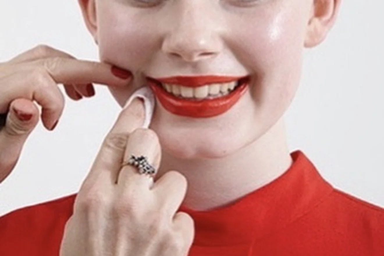 Close-up of a woman smiling with red lipstick, applying lipstick with her finger, wearing a red top, against a plain background.