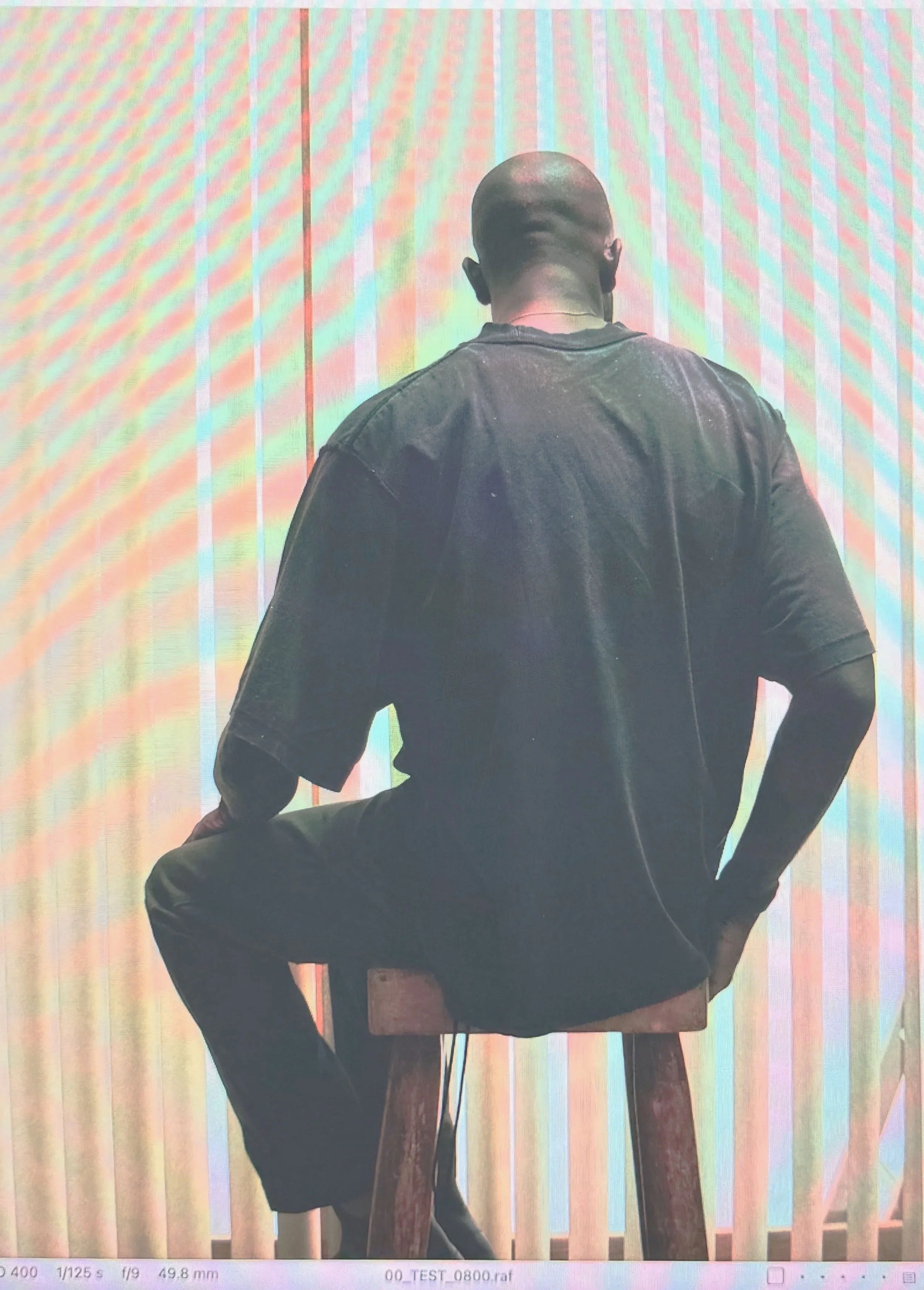 A person with dark skin, bald head, and wearing a black t-shirt sitting on a wooden stool, facing away from the camera against a colorful, art-like background.