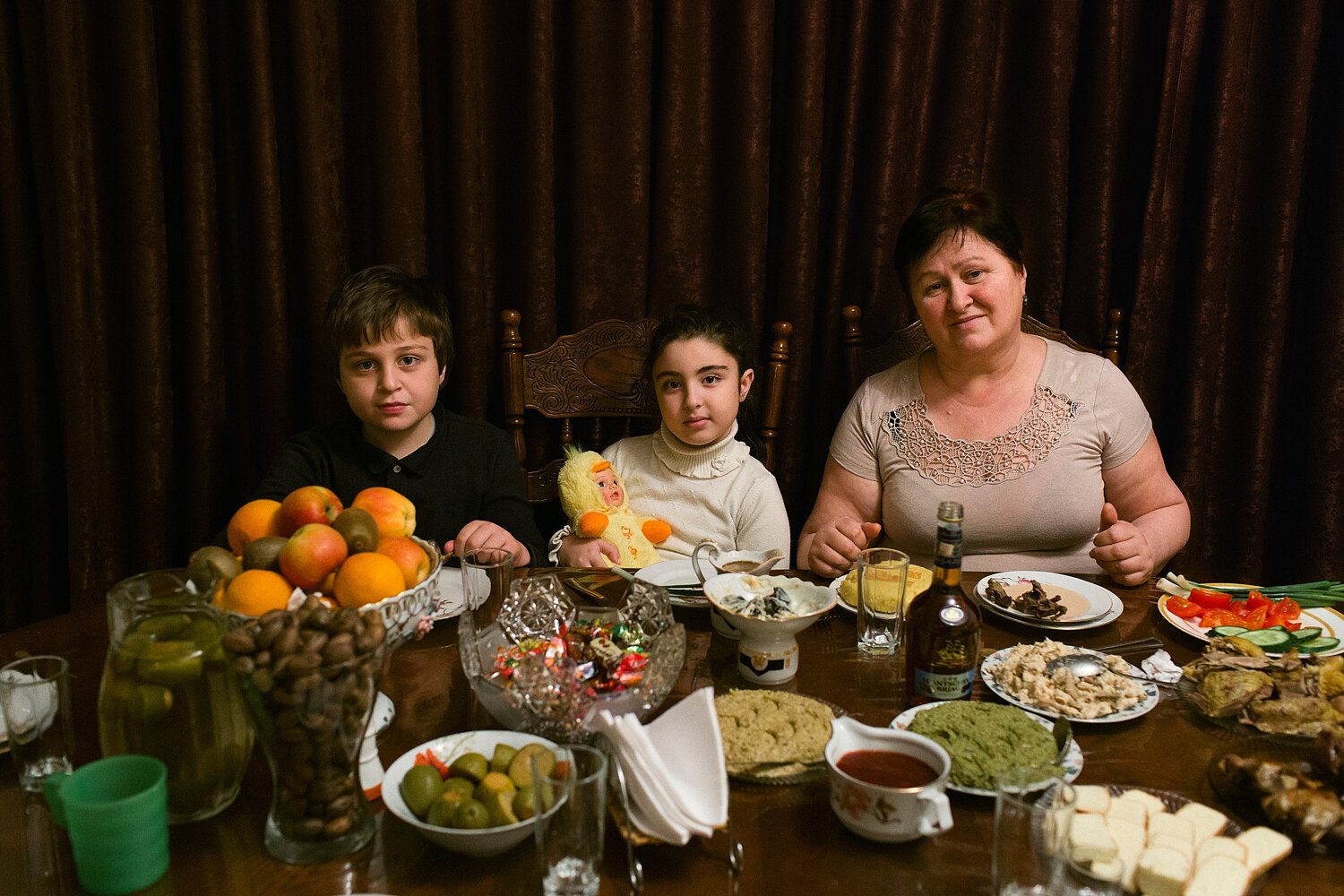 Abkhazia, Sukhum, 06/01/2016. Adlejba Rimma (55) with her grandchildren Sadya (7) and Sayat (9) in front of the table with traditional food, mamaliga (maize porridge), Ajika (spicy dip), Sulguni (cheese), vegetables and fruits - a typical family din