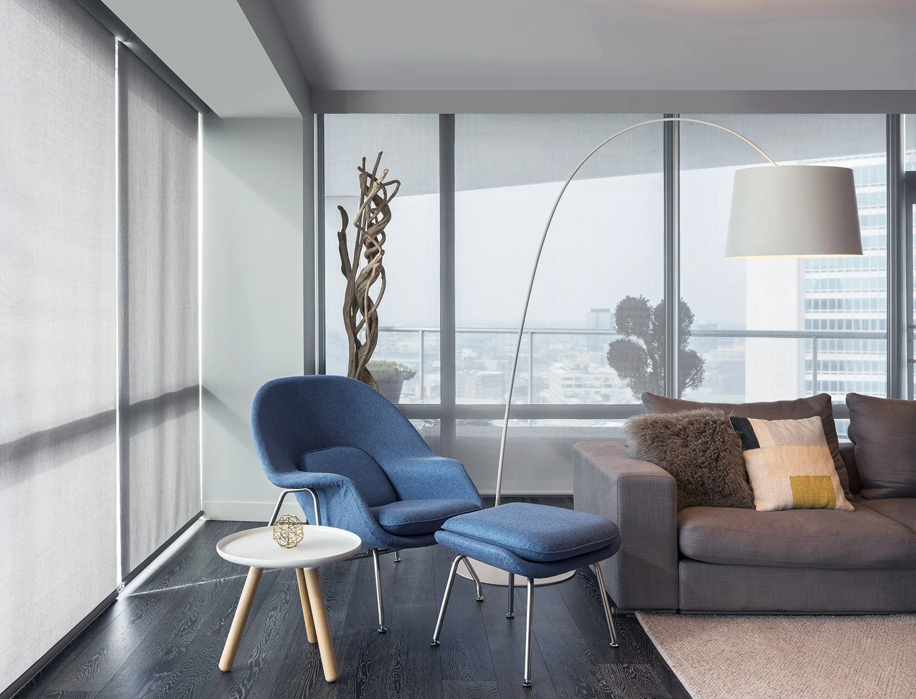 Modern living room with a gray sofa, blue armchair and footrest, a white side table with decorative object, tall lamp with curved stand, large windows with city view, sheer window coverings, and decorative plants.