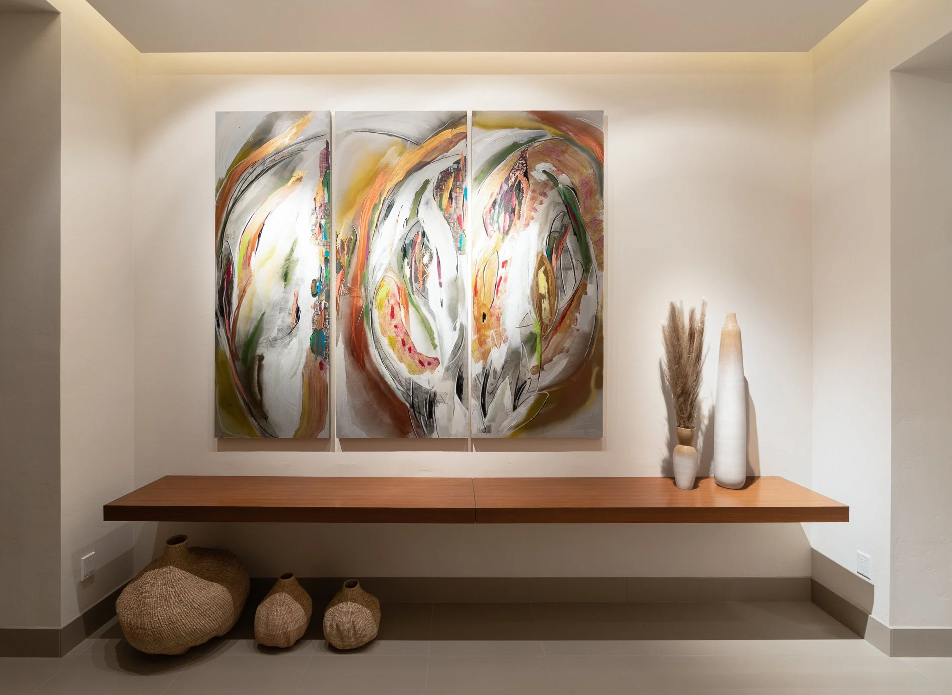 Interior wall with three large abstract paintings, a floating wooden shelf, and decorative vases and baskets underneath.