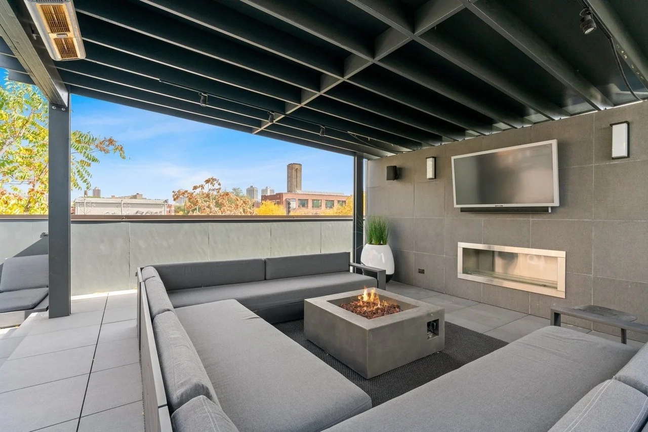 Outdoor rooftop lounge area with gray sectional sofa, a modern fire pit table, wall-mounted flat-screen TV, and city view in the background.