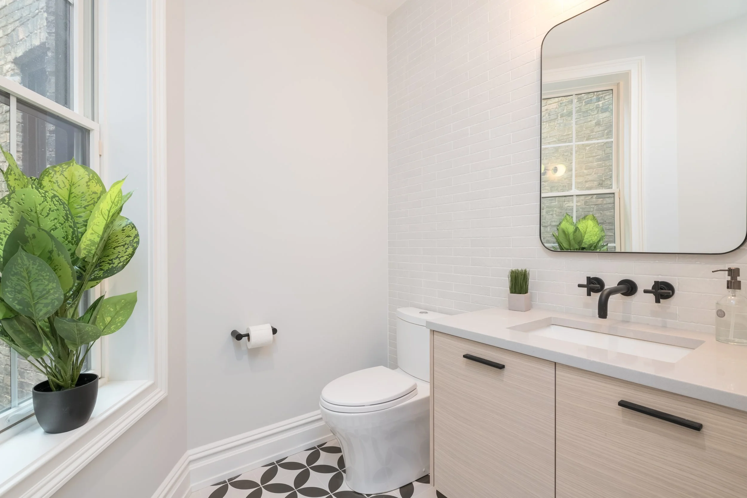 Modern bathroom with white walls, a round mirror, a white sink with black fixtures, a light wood vanity, a potted plant, a window with a plant on the sill, and patterned black and white floor tiles.