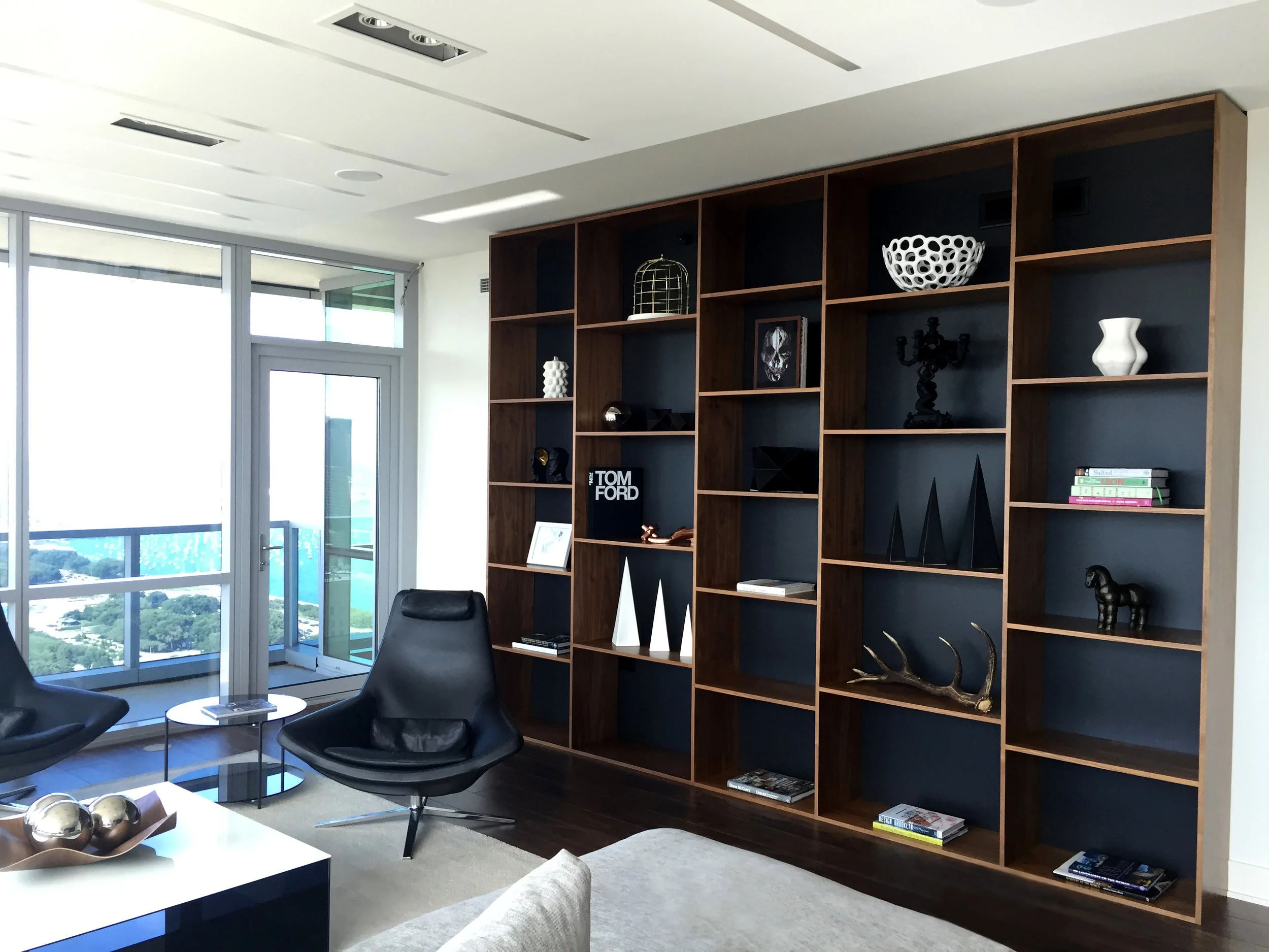 Modern living room with large window, black leather chairs, and a wooden bookshelf filled with decorative objects and books.