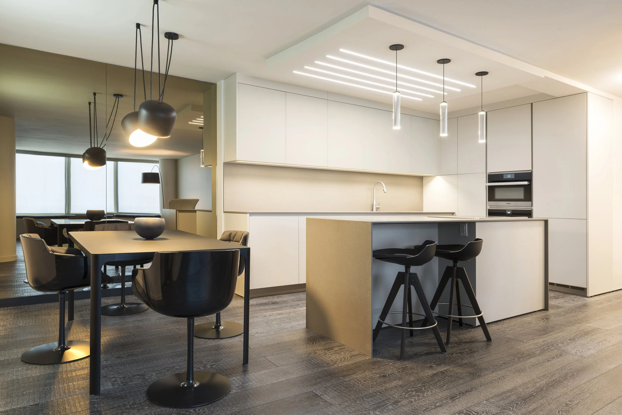 Modern kitchen and dining area with white cabinetry, black and beige bar stools, black dining chairs, and black pendant lights, with a wood floor and large windows in the background.