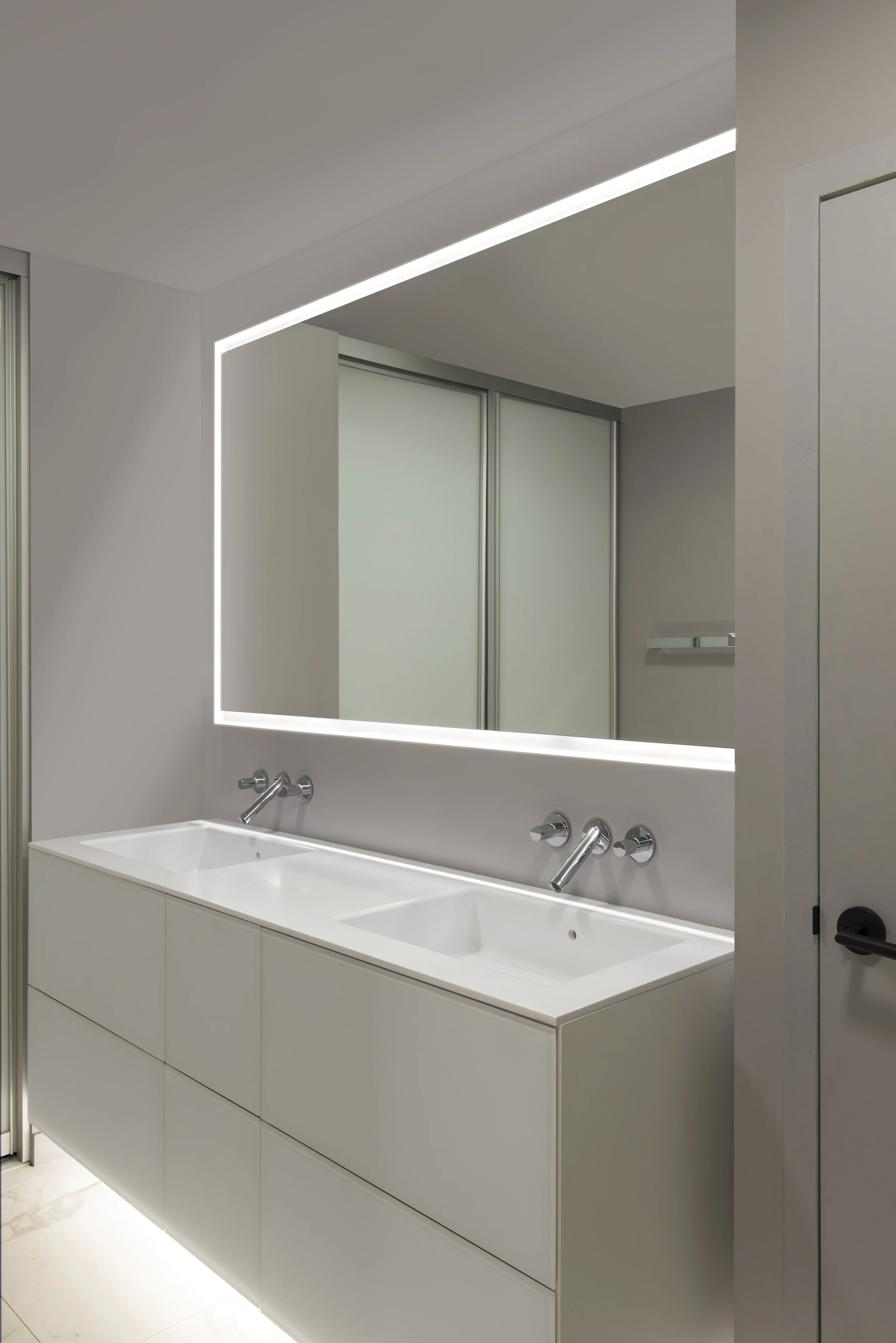 Modern bathroom with a double sink vanity, large illuminated mirror, and sliding closet doors in the background.