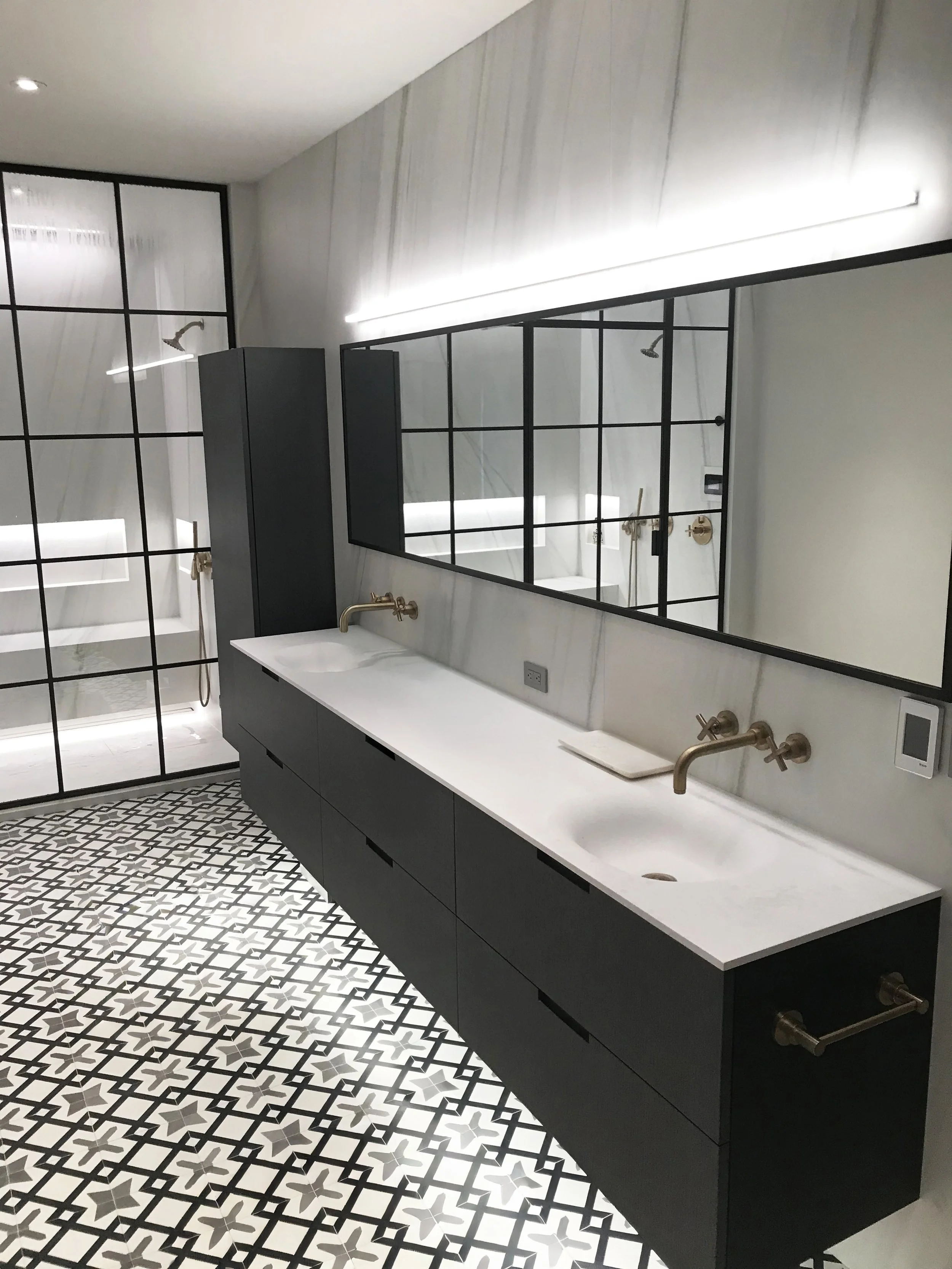 Modern bathroom with black and white patterned floor, white countertop with two sinks, gold faucets, large mirror, and glass shower enclosure with black framing.