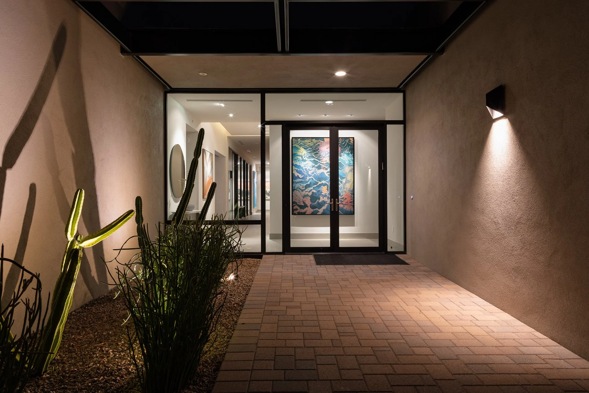 Night view of a building entrance with double glass doors, a large colorful wave art piece inside, desert plants on the left, and exterior wall lighting