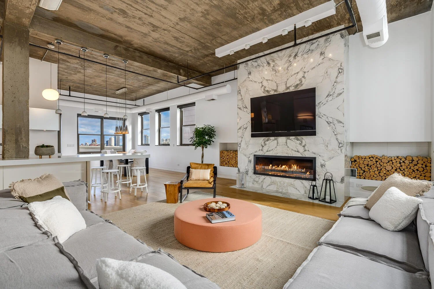 Modern apartment living room with gray sofas, a round pink ottoman, a marble fireplace, a flat-screen TV, and large windows with a city view.