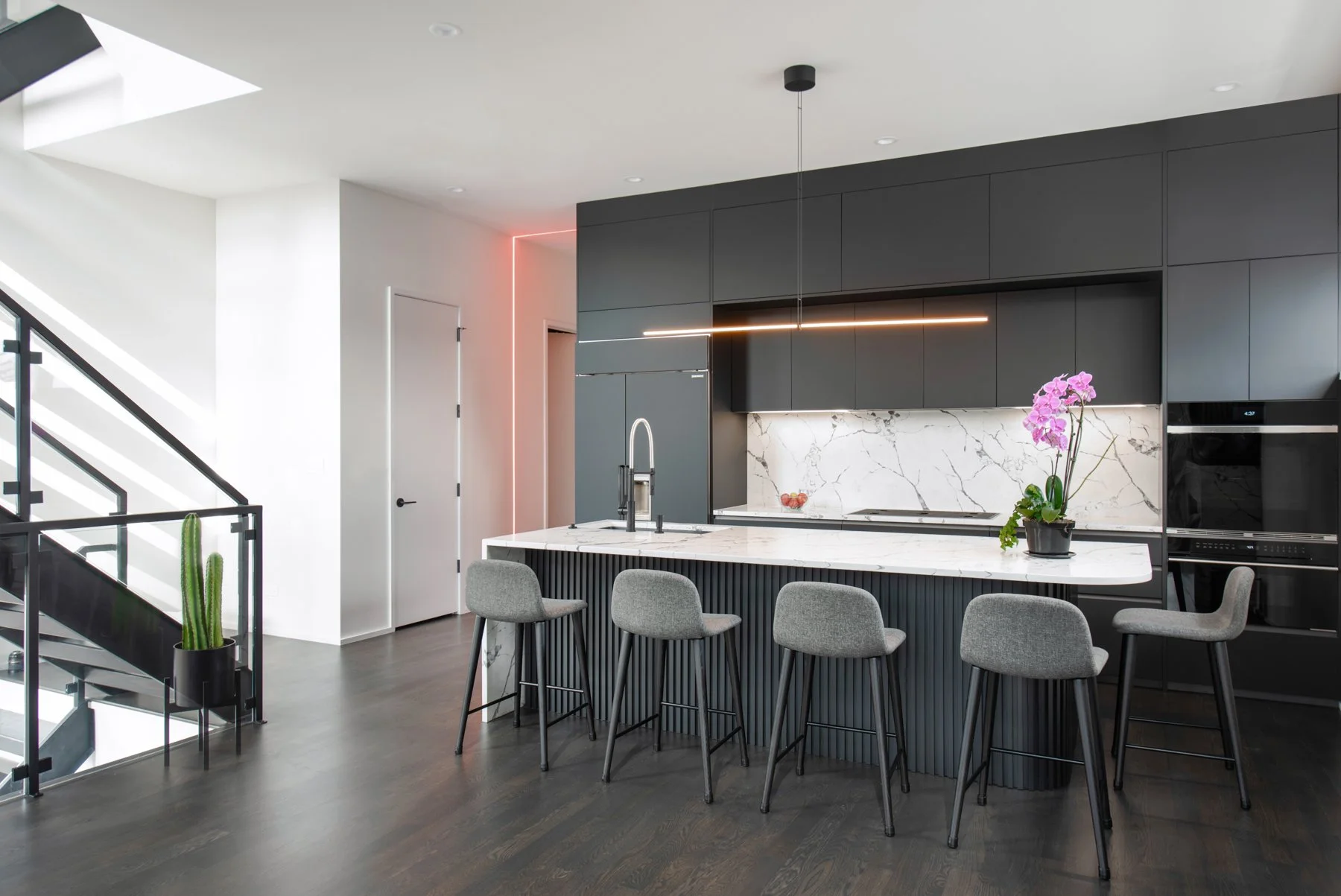 Modern kitchen with black cabinets, white marble backsplash and island, gray bar stools, pink orchid, cactus plant, and stairs with glass railing.