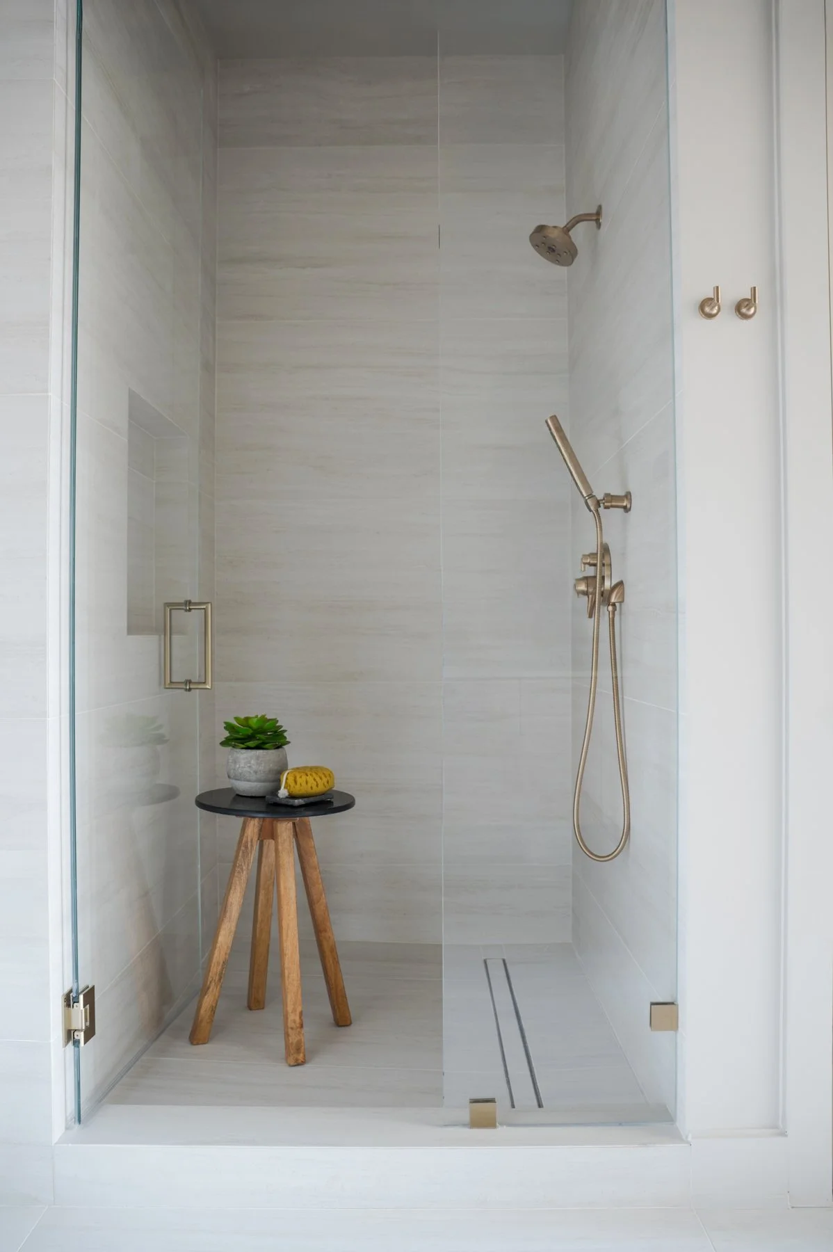 Empty modern glass-enclosed shower with beige tile walls, a silver showerhead, handheld shower, built-in shelf, two gold hooks, and a small wooden stool with a plant and sponge inside.