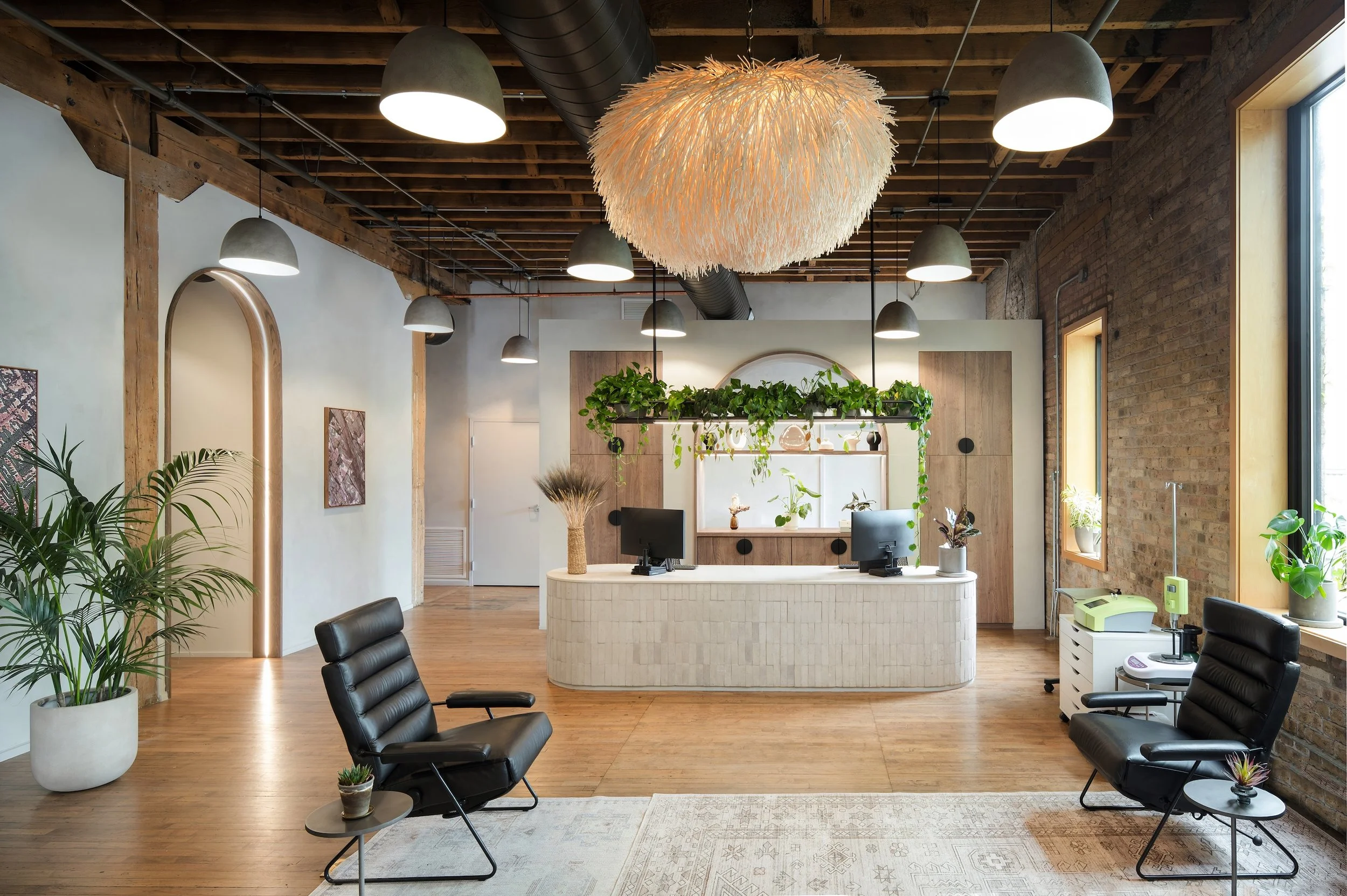 Modern office reception area with wood floors, a white curved reception desk with two computer monitors, hanging greenery, and seating area with black lounge chairs and small side tables, large windows, exposed brick and wooden beams ceiling, and dec