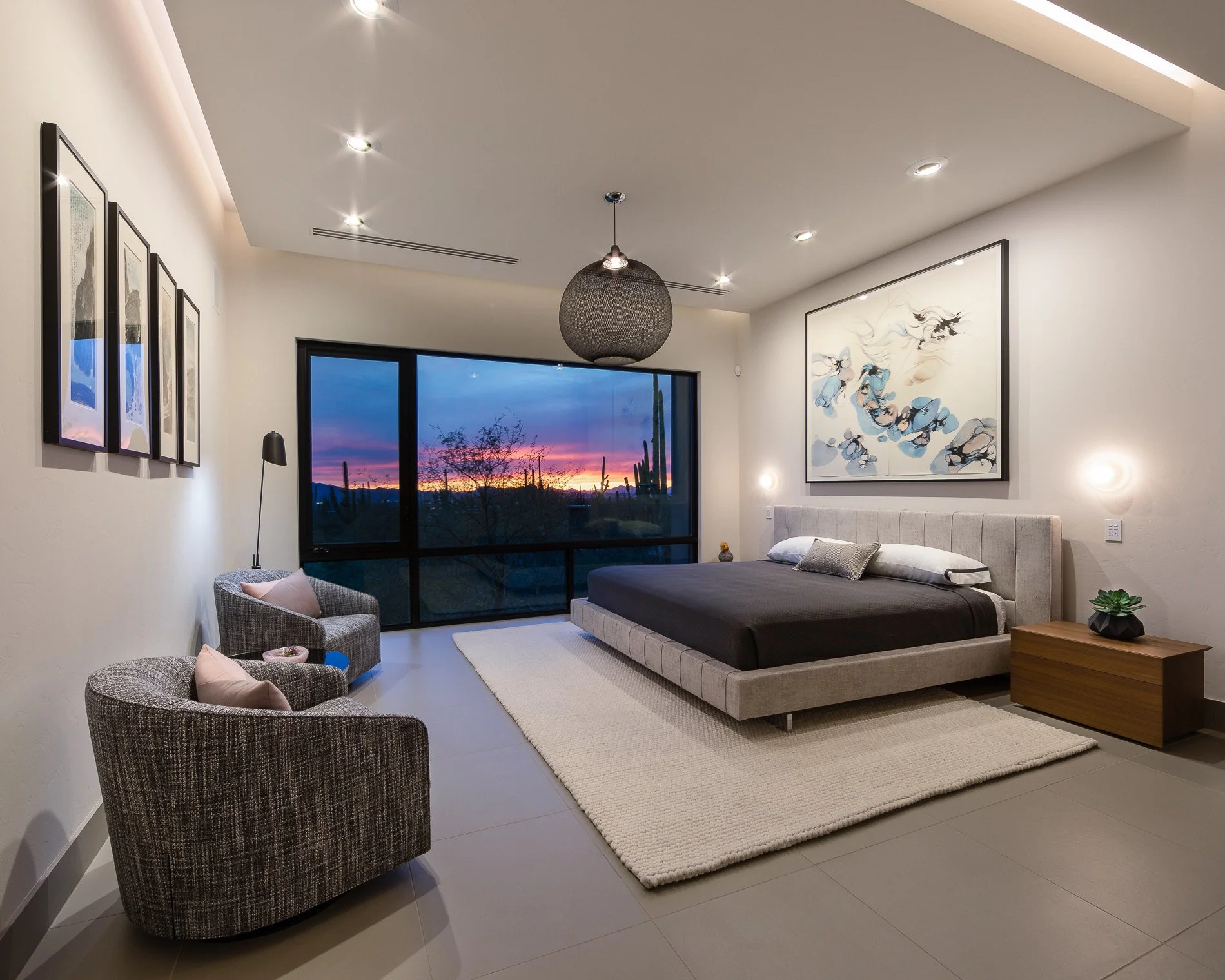 Modern bedroom with large window showing a sunset and desert landscape with cacti, decorated with abstract artwork and minimal furniture, including a bed, nightstand, armchairs, and a rug.