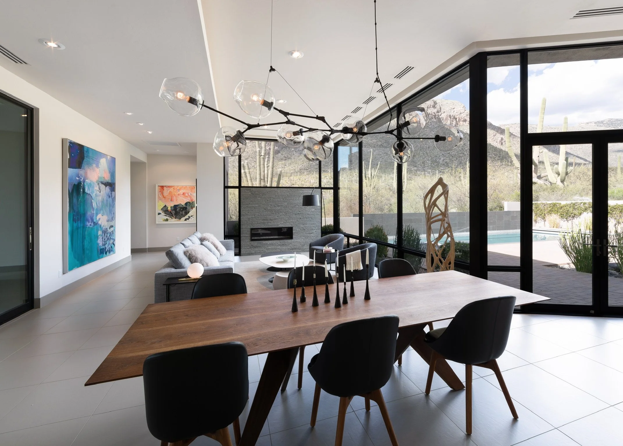 Modern living and dining room with large floor-to-ceiling windows showing desert landscape, cactus, and mountains outside. Interior features a wooden dining table with six black chairs, a sitting area with a gray sofa, abstract art on the walls, a fi