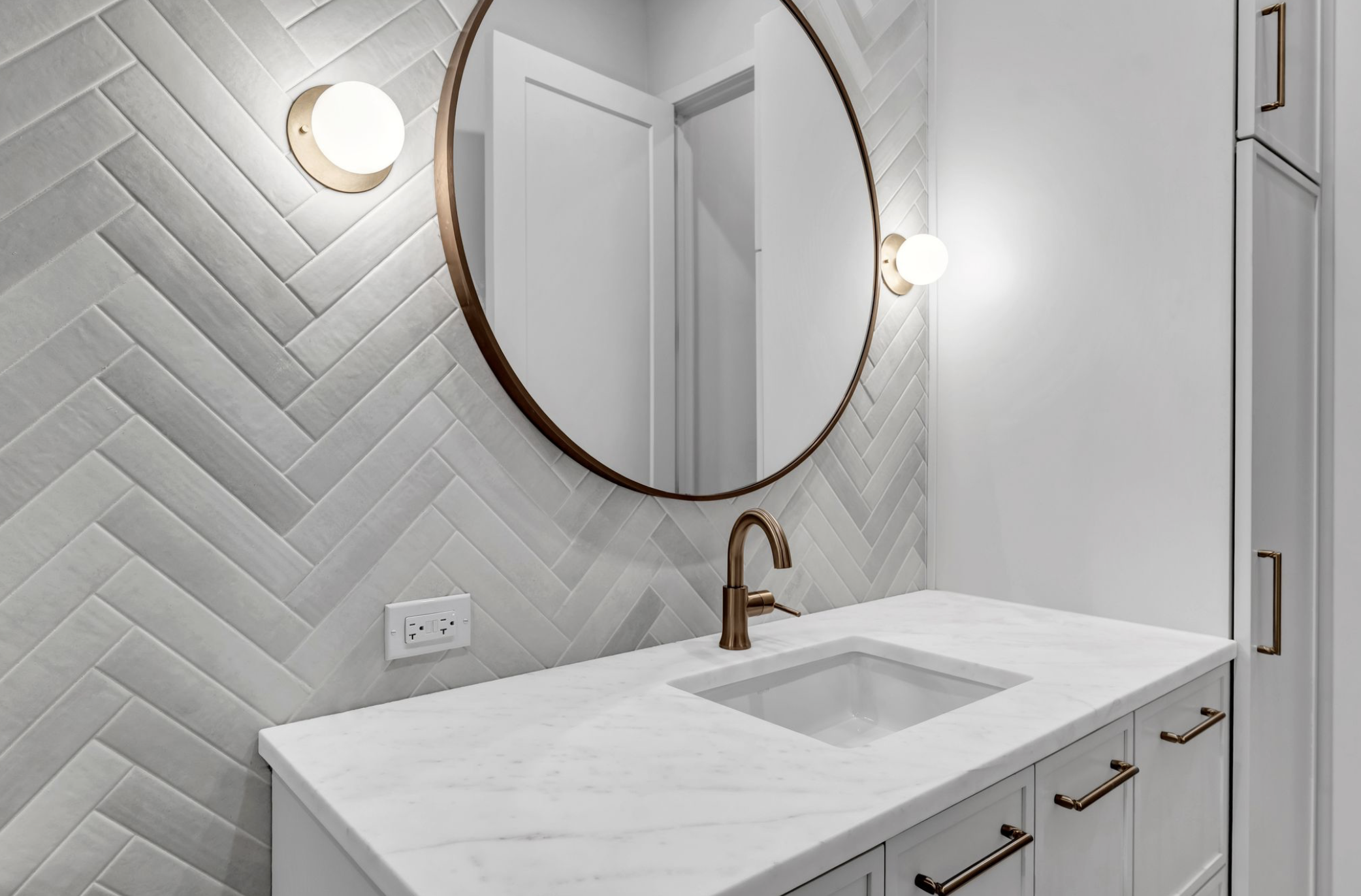 Modern bathroom vanity with a white marble countertop, a built-in sink, a bronze faucet, an oval mirror, and wall-mounted lights with round white shades. The wall behind the vanity features a textured, herringbone-patterned tile, and there are white 
