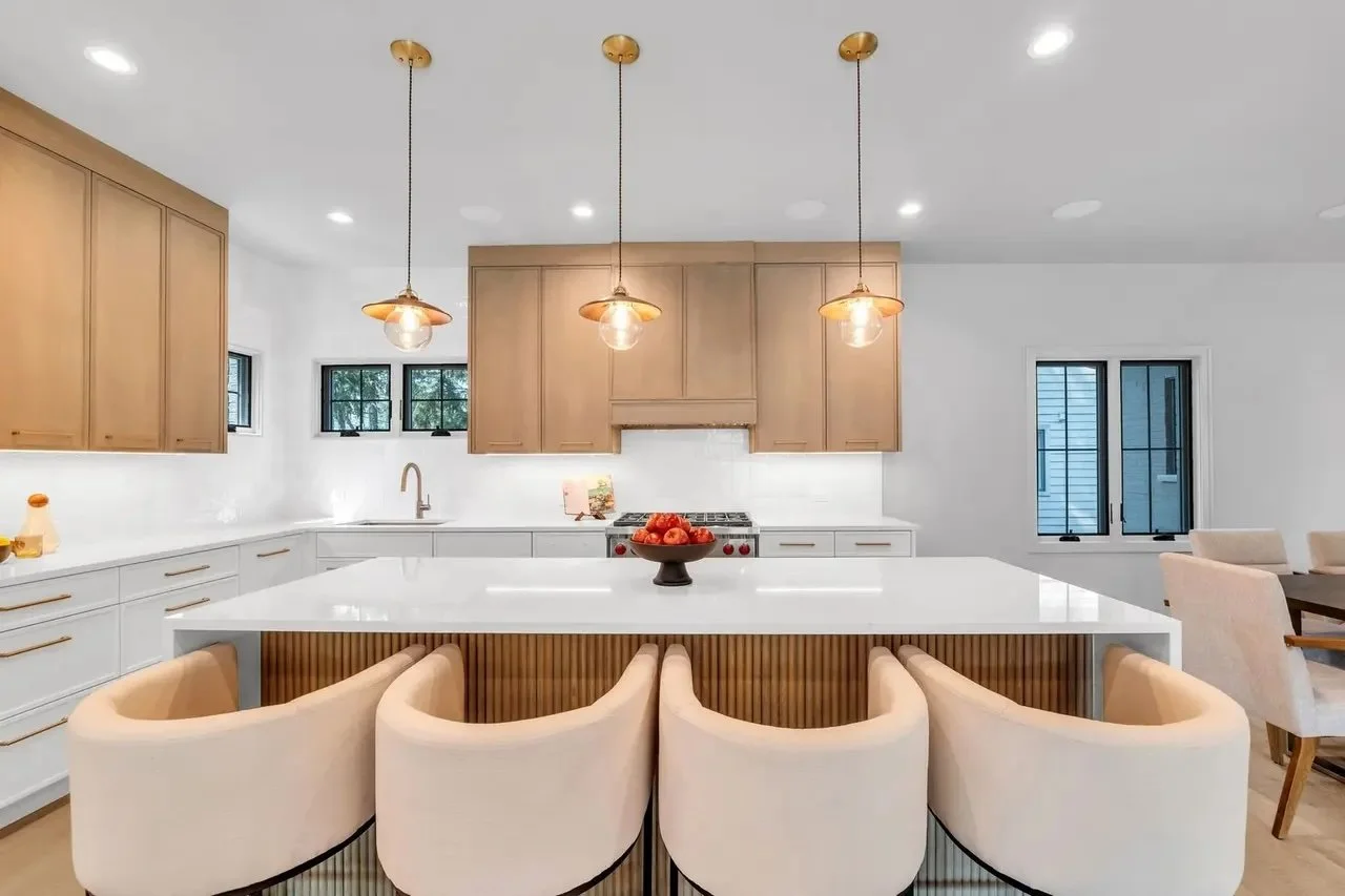 Modern kitchen with beige cabinetry, white countertops, and a large island with six beige upholstered chairs. Pendant lights hang over the island. There are three small windows above the sink and two larger windows to the right, with a dining area pa