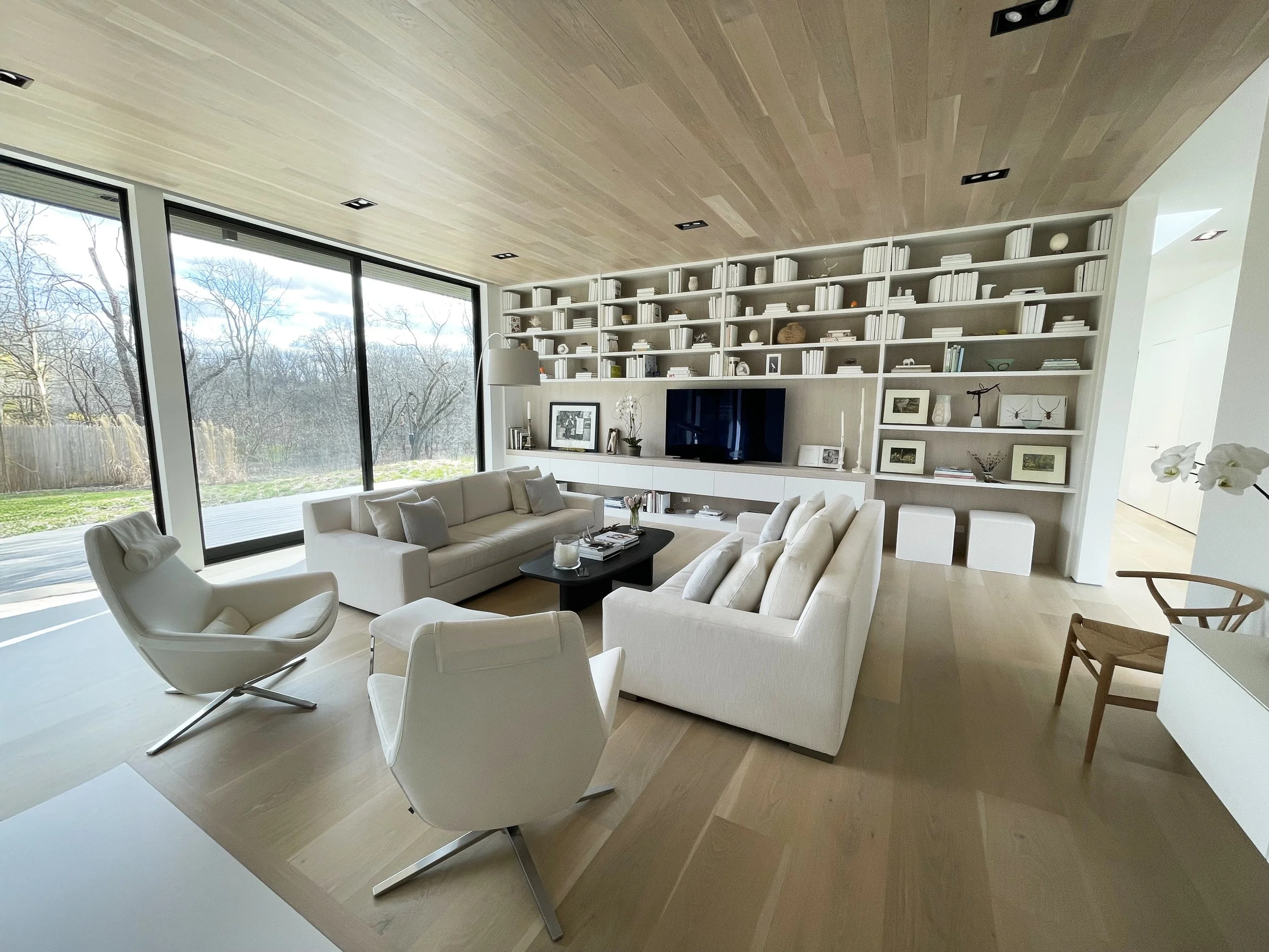Modern living room with large floor-to-ceiling windows, white sofas, armchairs, a black coffee table, and a built-in bookshelf with a wall-mounted TV.