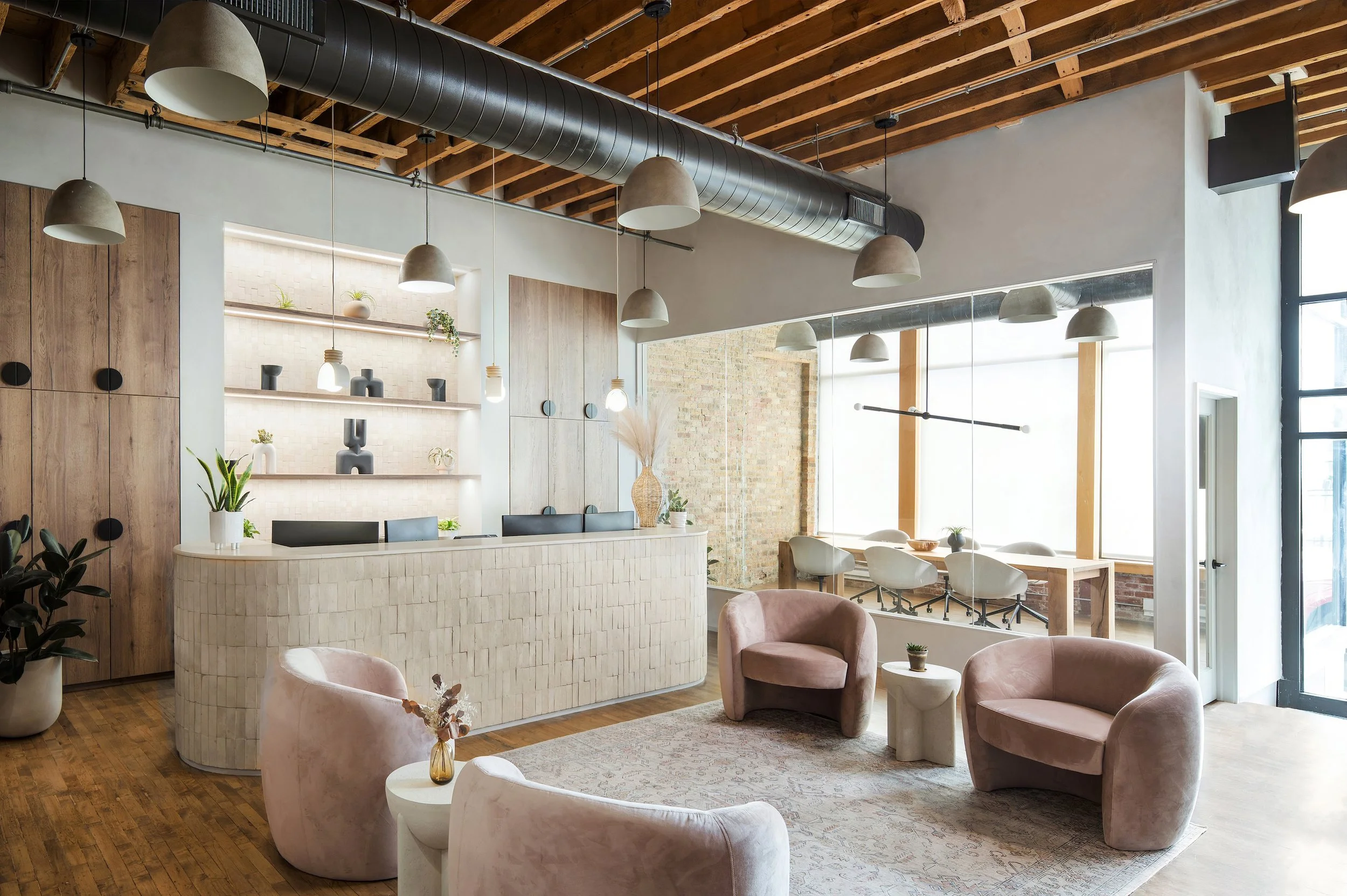 Modern, minimalist reception area with curved white brick front desk, three plush pink chairs, white side tables, potted plants, and large windows allowing natural light, with wooden beams and ceiling ductwork visible.