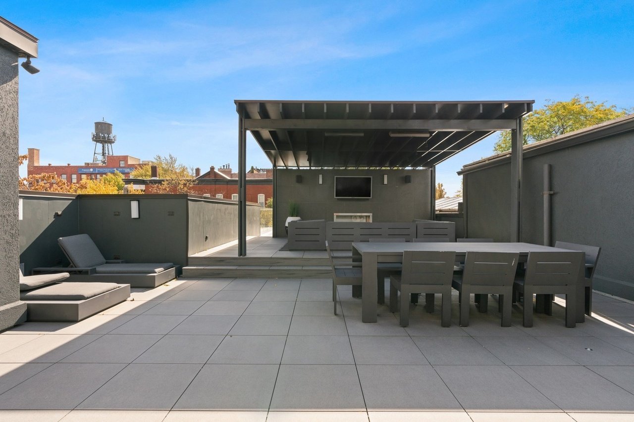 Modern rooftop patio with outdoor furniture, a covered dining area, lounge chairs, and a wall-mounted television under a metal pergola, with a cityscape and blue sky in the background.