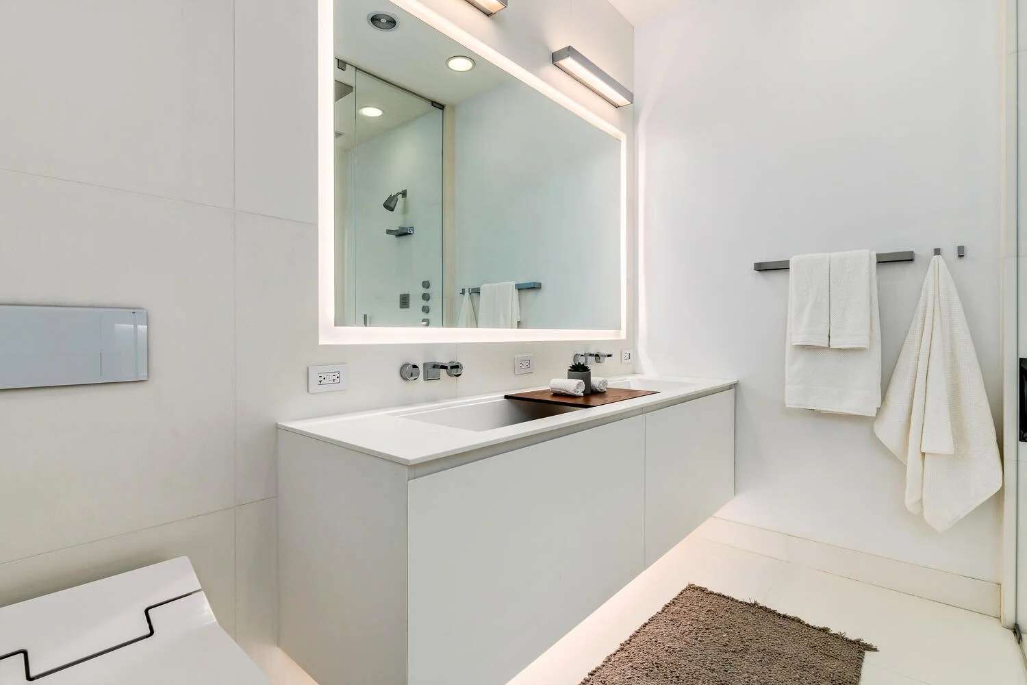 Modern minimalist bathroom with white walls and a large backlit mirror above the white vanity, which features a built-in sink, black faucet, and a small green plant with towels on a wooden tray. Towel bars with white towels are mounted on the wall, a