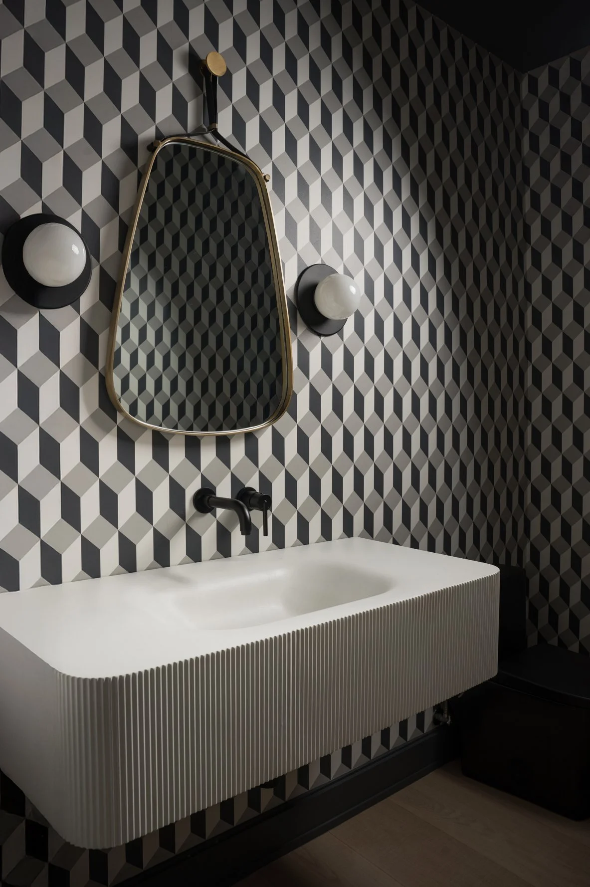 Modern bathroom with geometric black, white, and gray patterned wallpaper, a white textured sink, an oval mirror with a gold frame, and two round white wall sconces.