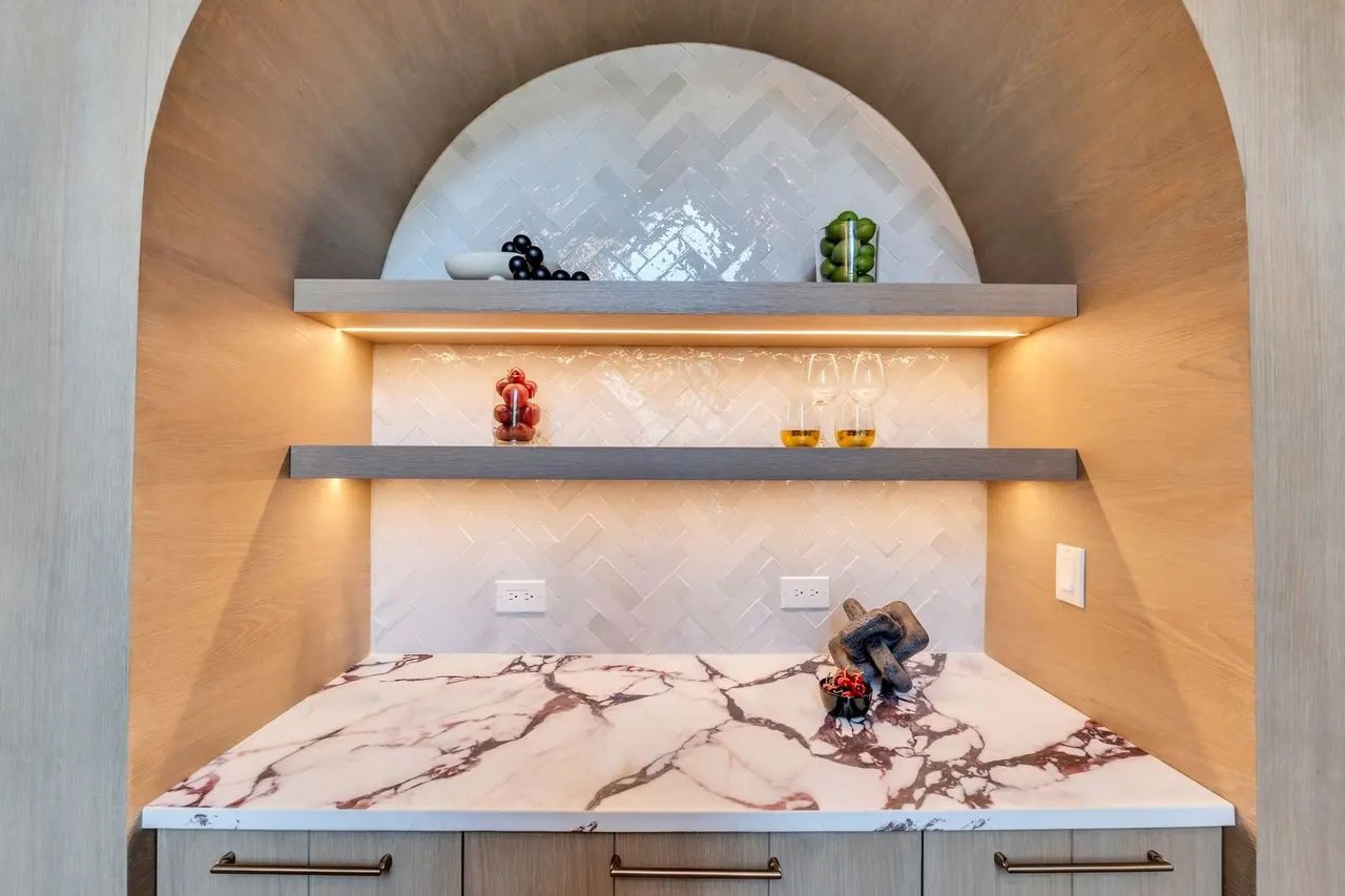 A kitchen corner with two gray floating shelves above a marble countertop. The shelves hold decorative jars with blackberries, limes, and red berries. The backsplash behind the shelves has a white herringbone tile pattern. There are electrical outlet