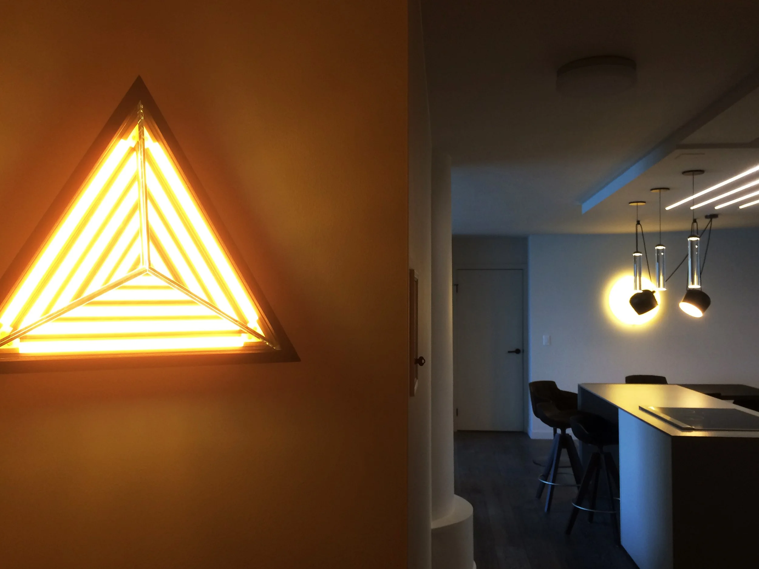 Interior view of a modern kitchen and dining area with various lighting fixtures, including a triangular neon wall lamp and three hanging spotlights over the counter.
