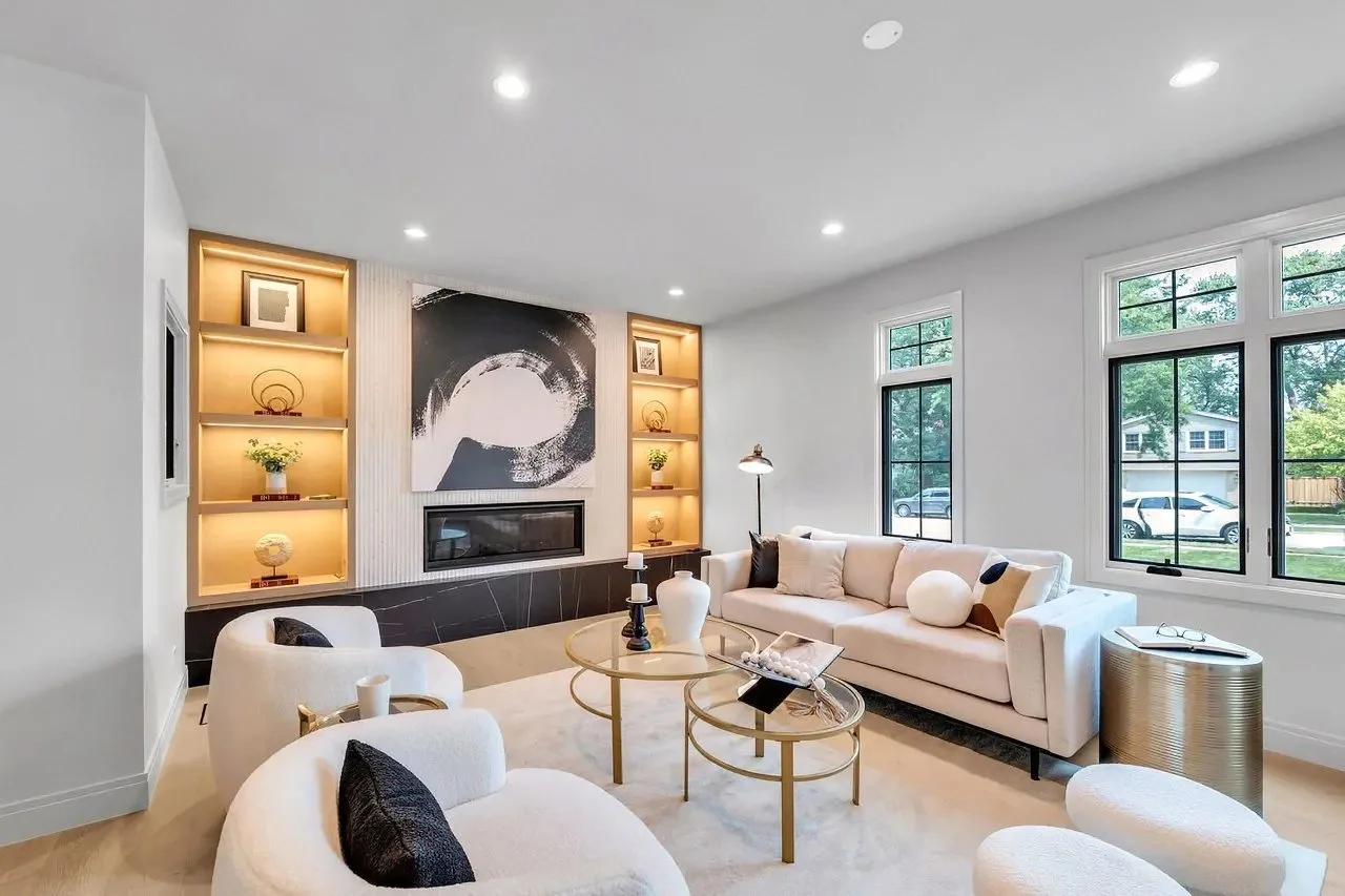 A modern living room with white walls, large windows, a light-colored sofa with black and white pillows, matching armchairs, a round side table, and decorative items on built-in shelves with warm lighting.