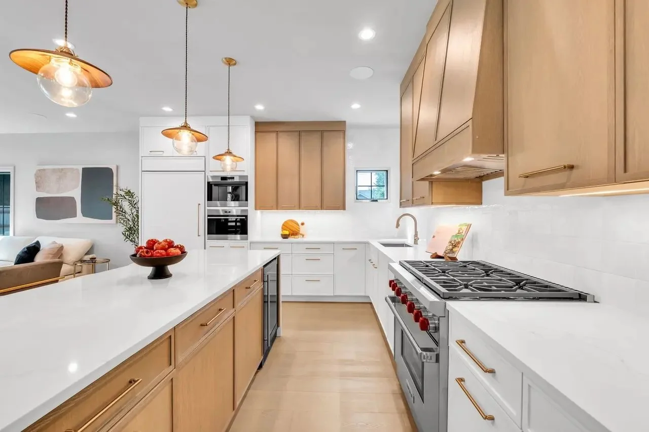 Modern kitchen with white countertops, light wood cabinets, a stainless steel oven, and a gas cooktop; decorative lighting fixtures hanging from the ceiling; bowl of red apples on the counter.