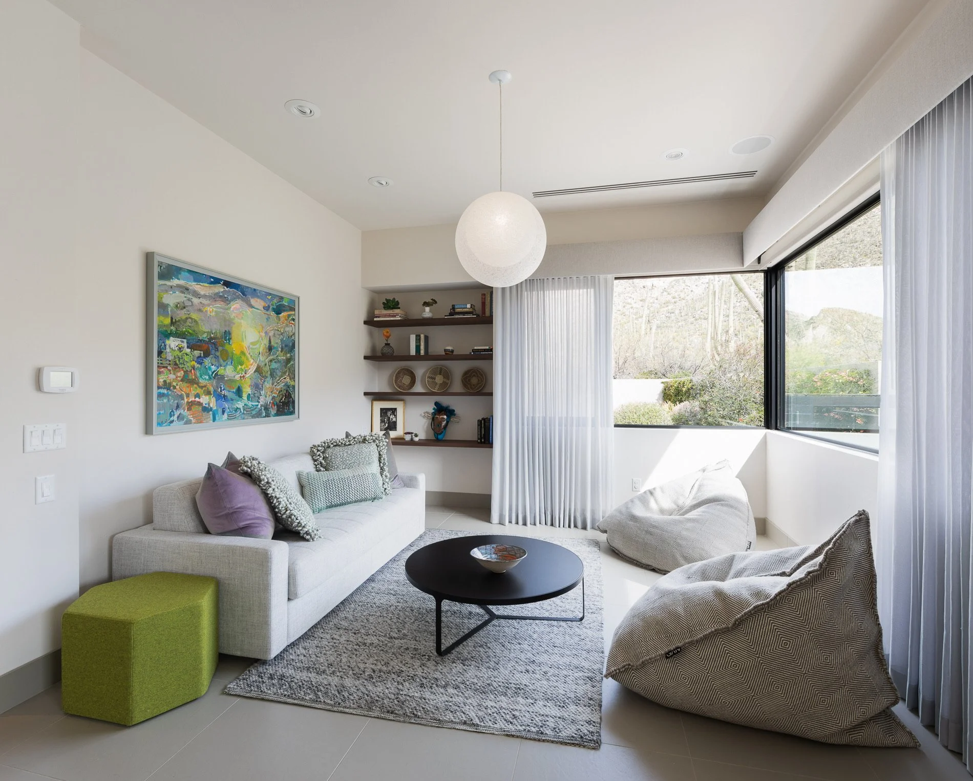 Modern living room with a white sofa, colorful abstract painting, open bookshelves, large windows with white curtains, and bean bag chairs.