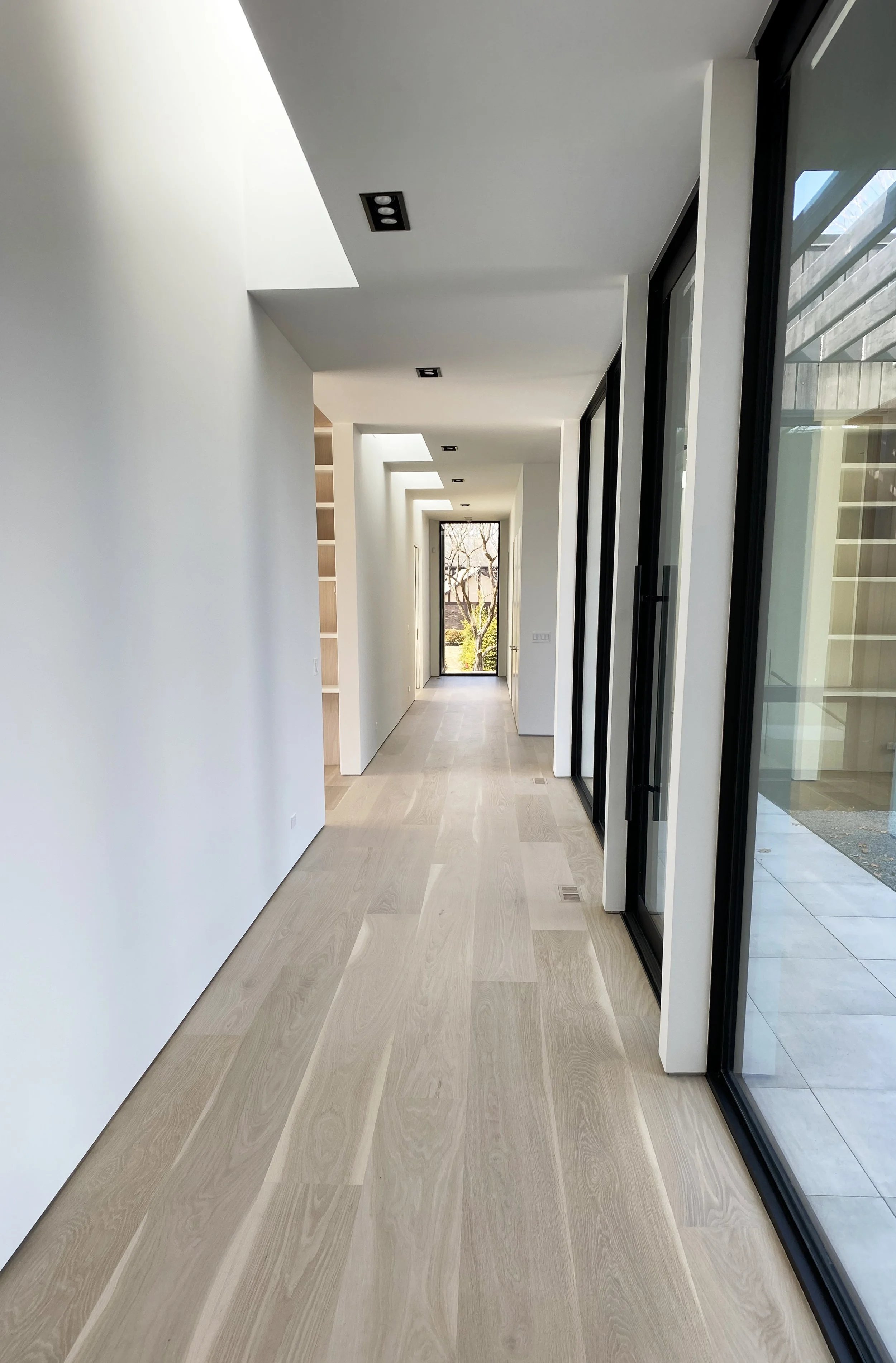A modern, minimalist hallway with light-colored wooden floors, white walls, ceiling lights, and large glass sliding doors on the right leading outside. At the end of the hallway, there is a view of trees and sunlight.