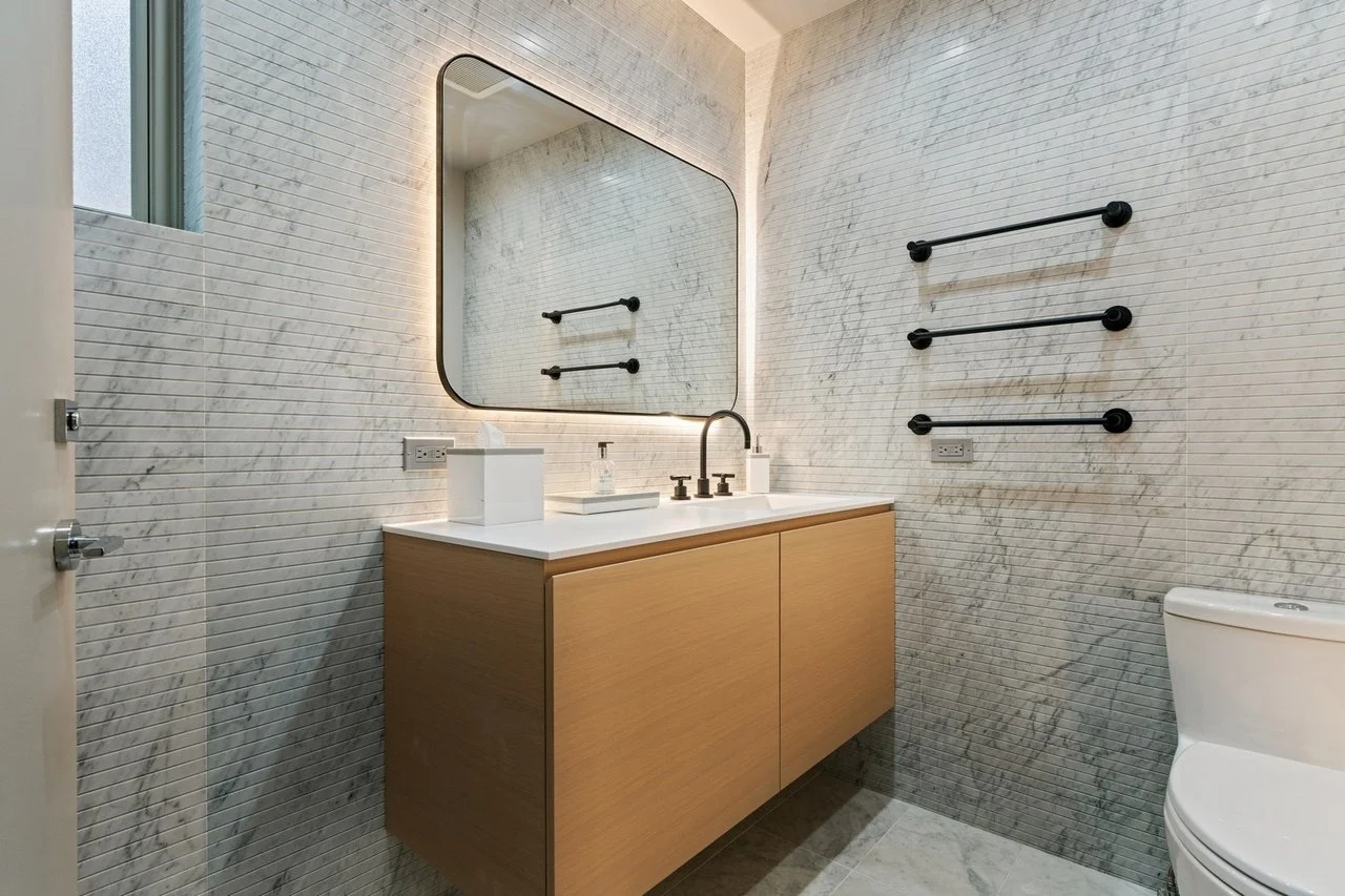 Modern bathroom with a large mirror, a wooden vanity with a white countertop, a sink with a black faucet, paper towels, and black towel racks on textured gray and white walls.