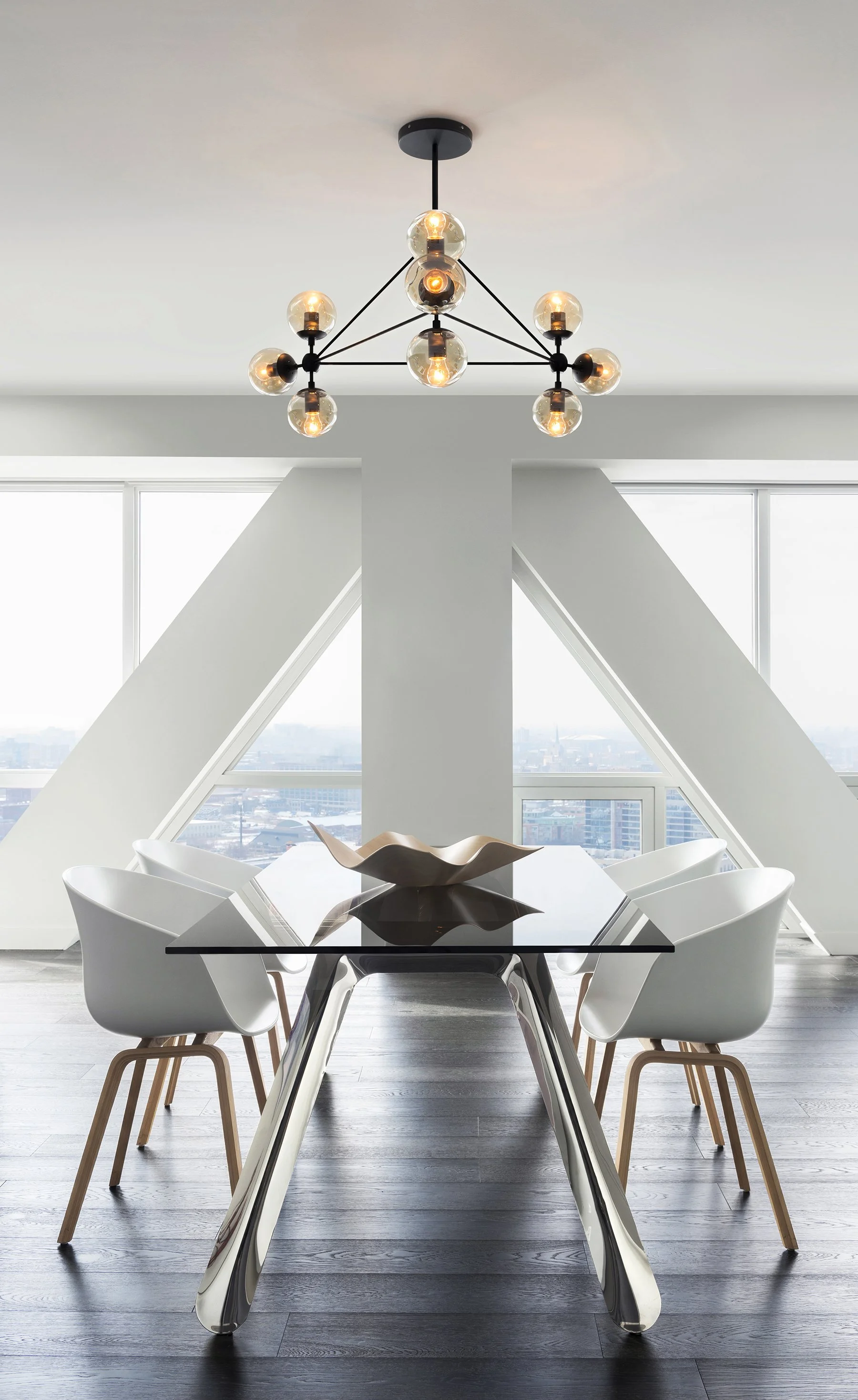 Modern dining room with a glass table, white chairs, a chandelier, and large windows with a city view.