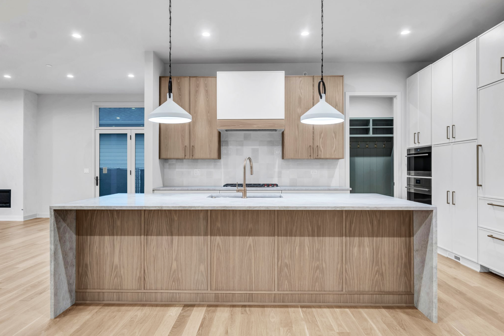 Modern kitchen with a large white island, wooden lower cabinets, and white upper cabinets, with pendant lights, a sink, and built-in appliances.