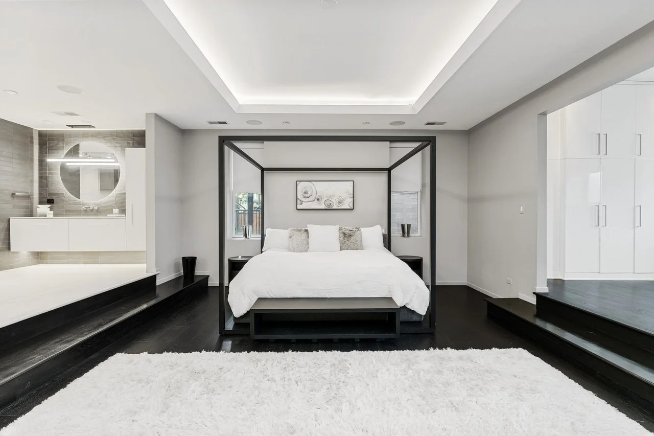 Modern bedroom with a canopy bed, white bedding, and decorative pillows, black nightstands, a textured white rug, and a bathroom in the background.