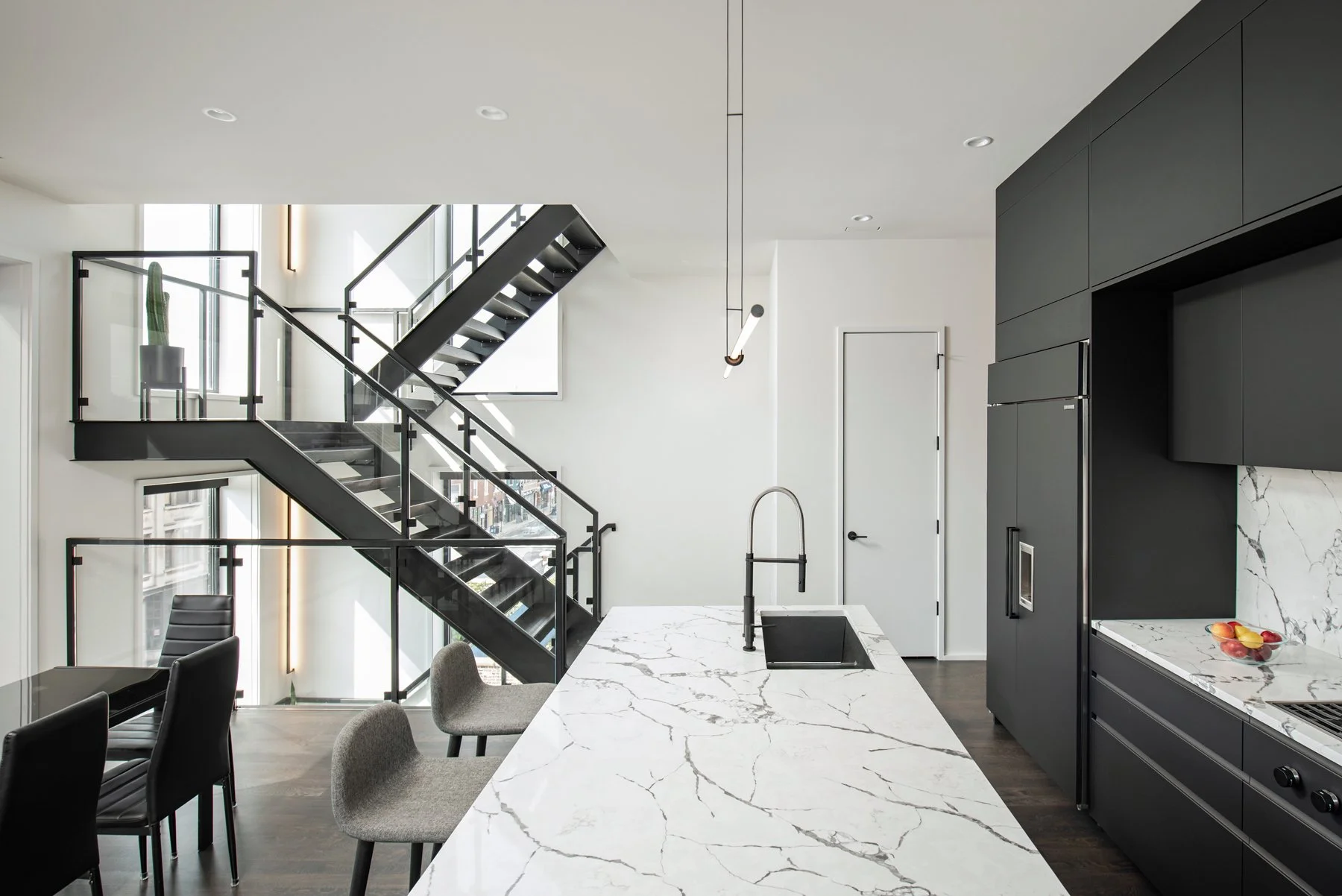 Modern kitchen with white marble countertop, black cabinetry, and a staircase with black metal railings leading to an upper floor.