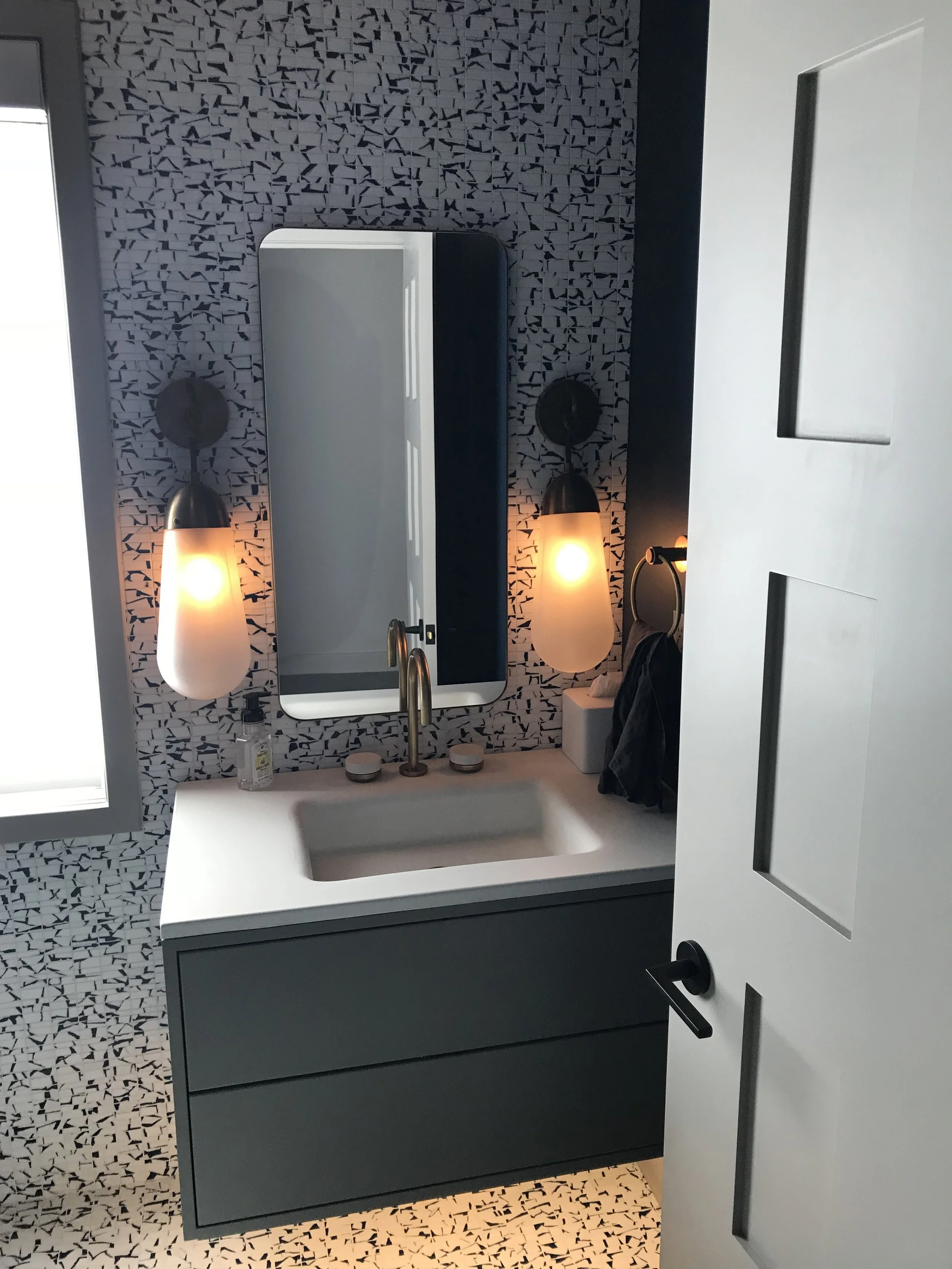 Modern bathroom with a black and white mosaic tile wall, a white vanity with a sink, a mirror, two black wall-mounted light fixtures with frosted glass shades, a small bottle of hand soap, a tissue box, and a hanging towel.