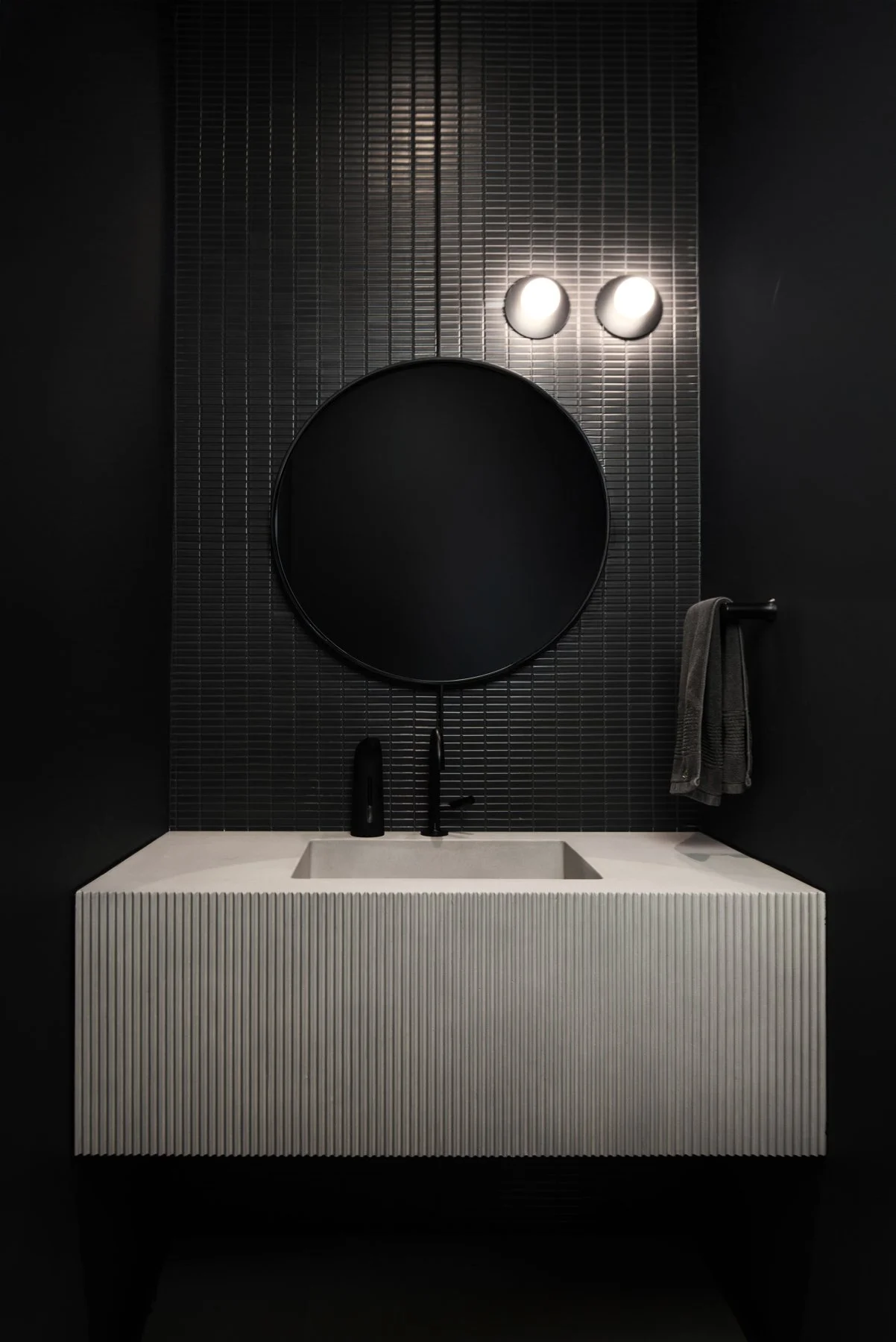 Modern powder room  with black mosaic tiled wall, round black mirror, white textured concrete sink, black faucet, black soap dispenser, black towel on a black towel bar, and two white wall-mounted lights.