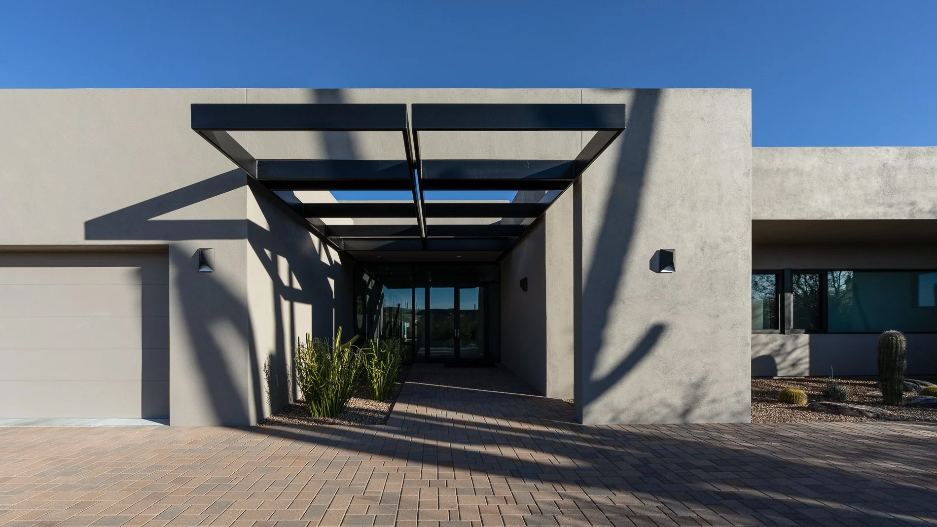 Modern house with flat roof, concrete walls, and a glass entrance. Shadows cast by nearby trees are visible on the wall, with desert plants in the landscaping.