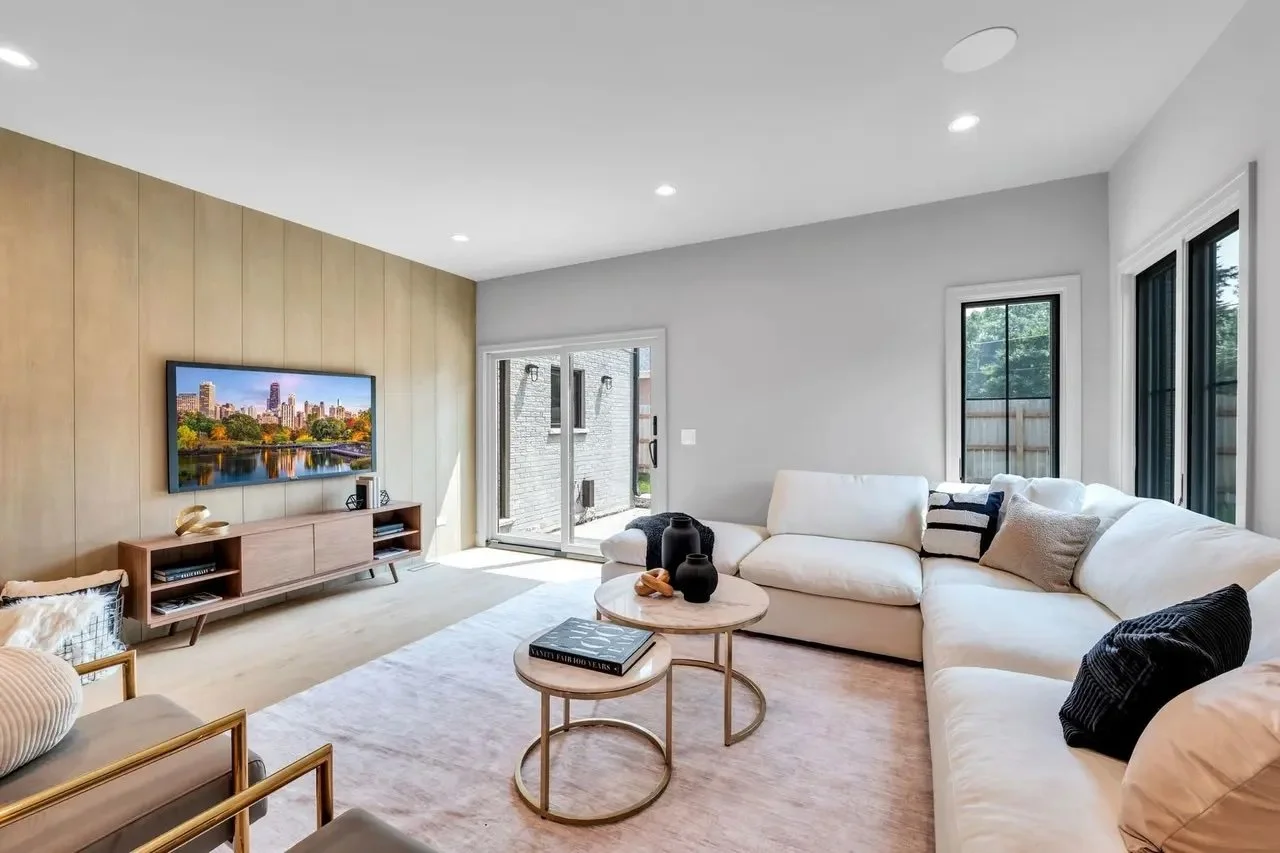 Modern living room with a large white sectional sofa, round coffee tables, a wall-mounted flat-screen TV, and large windows letting in natural light.