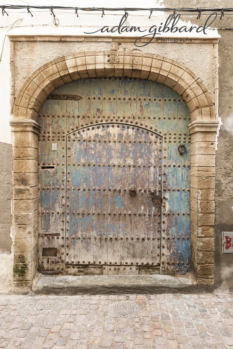 Old, weathered wooden door with rusted rivets, set in a stone archway, on a cobblestone street. Above it is a sign that reads 'adam gibbard photography'.