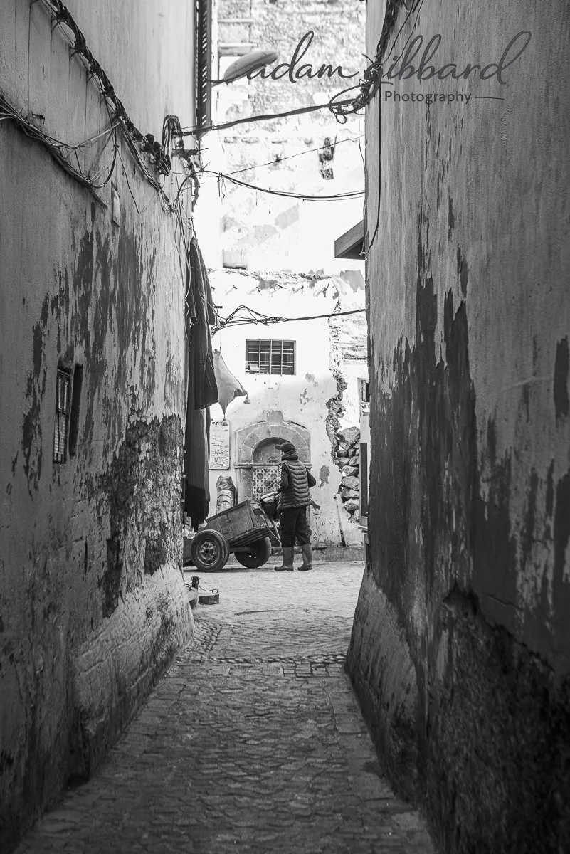 A narrow alleyway with worn, peeling walls leading to a courtyard where a person with a cart is visible.