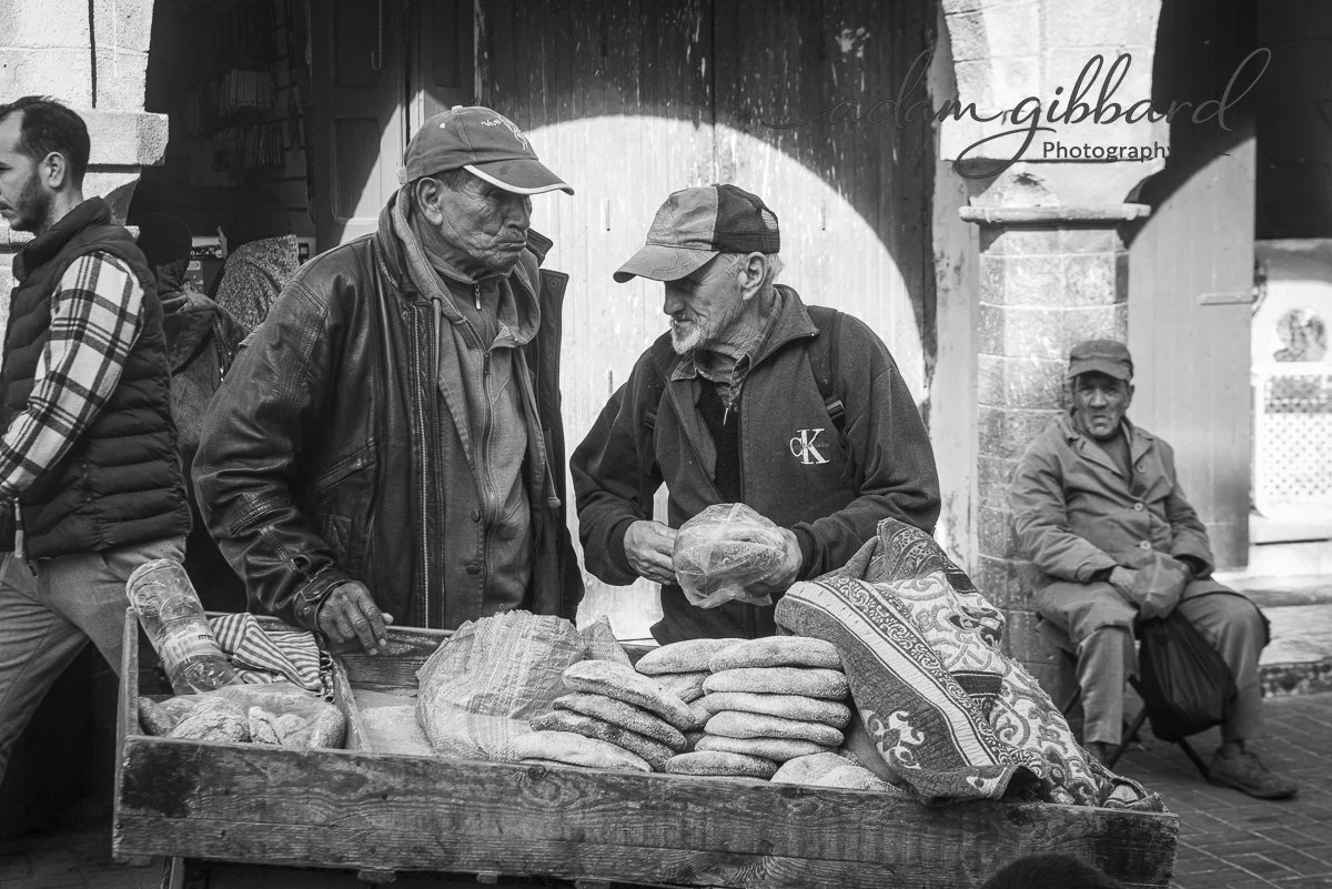 Two elderly men in jackets and caps buy bread from a street vendor. The vendor's cart has several loaves of bread and a patterned cloth. In the background, a man is sitting against a wall, and other people are walking nearby.