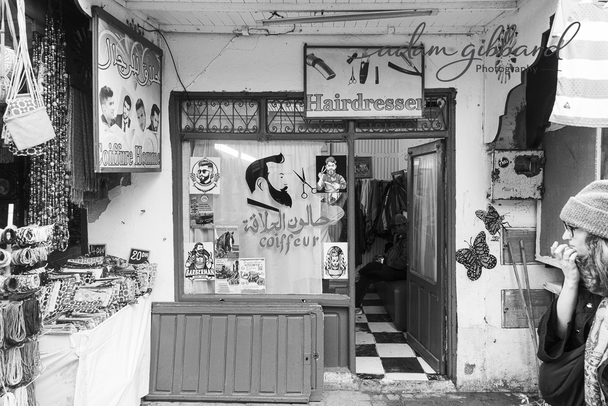 Street view of a small barbershop with a sign in Arabic and French that reads 'coiffeur,' with images of a bearded man and barber tools, and a nearby woman with a hat talking on her phone.