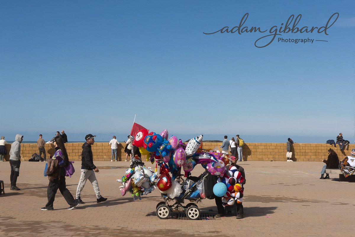 Street vendor selling colorful balloons near a seaside promenade with people walking and sitting, and a clear blue sky in the background.