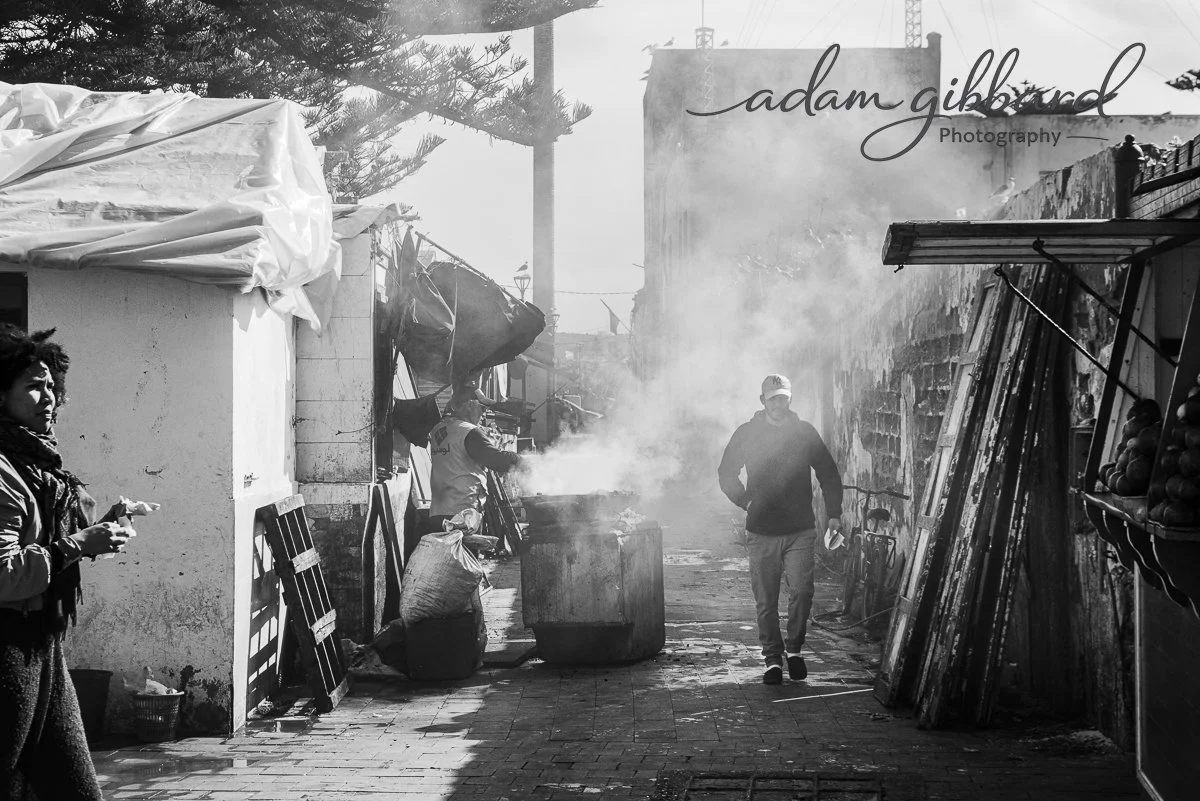 A black and white photo of an outdoor market street with people walking. Smoke rises from a large container in the center. To the left, a woman stands with a snack. To the right, a man in a hoodie and cap walks. Market stalls with wood planks and a f