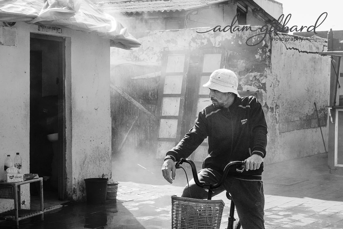 A man wearing a bucket hat and a black jacket rides a bicycle in front of a weathered building, with sunlight and some dust or mist in the air.