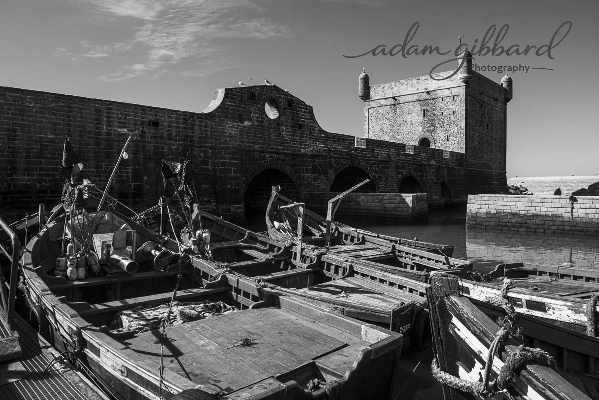 Black and white photo of abandoned boats docked near an old stone fortress or castle by the water, with seagulls perched on the structure.