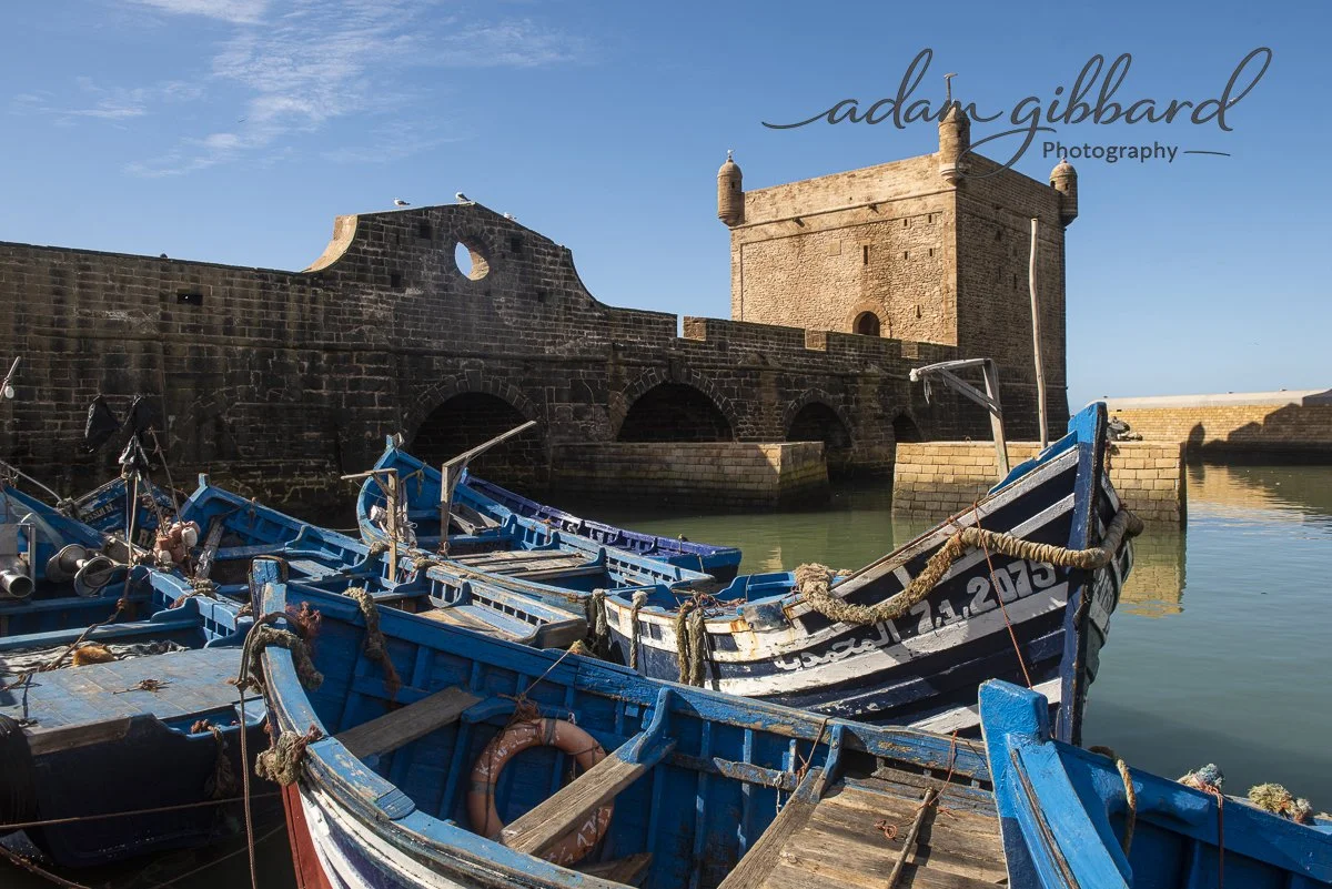A harbor with several blue boats docked in front of a historic stone fortress by the water, under a clear blue sky.